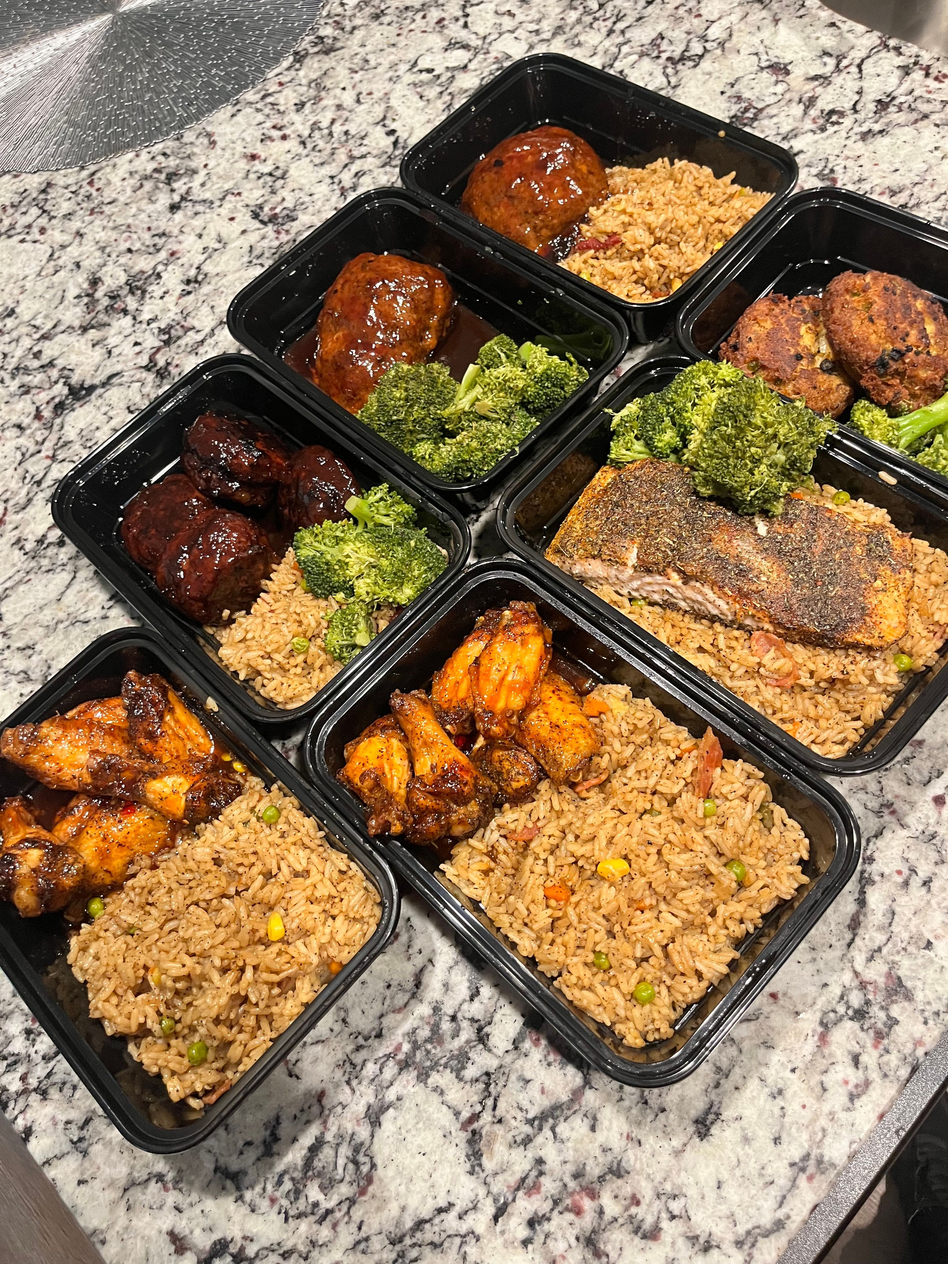 Four meal containers with rice, grilled chicken, broccoli, and meatballs on a marble surface.