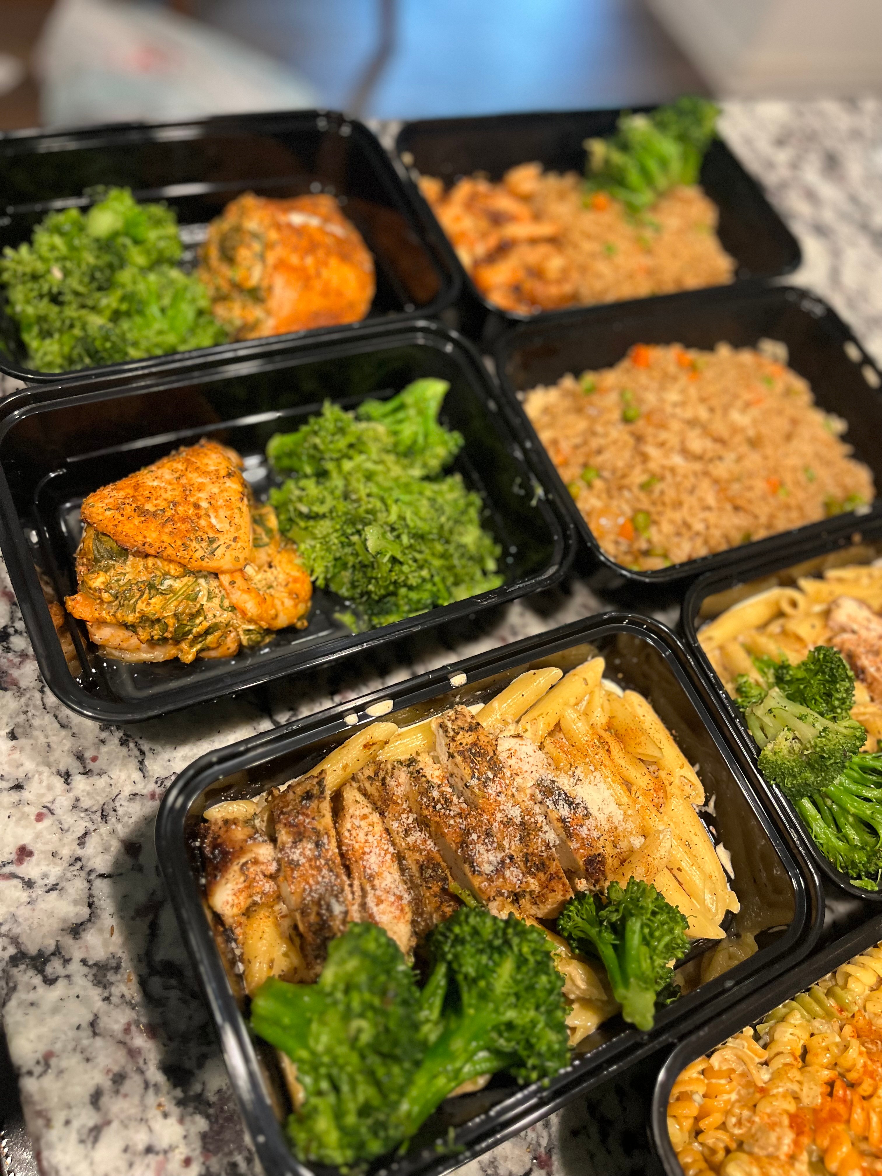Multiple black meal containers with rice, vegetables, and cooked dishes on a speckled surface.