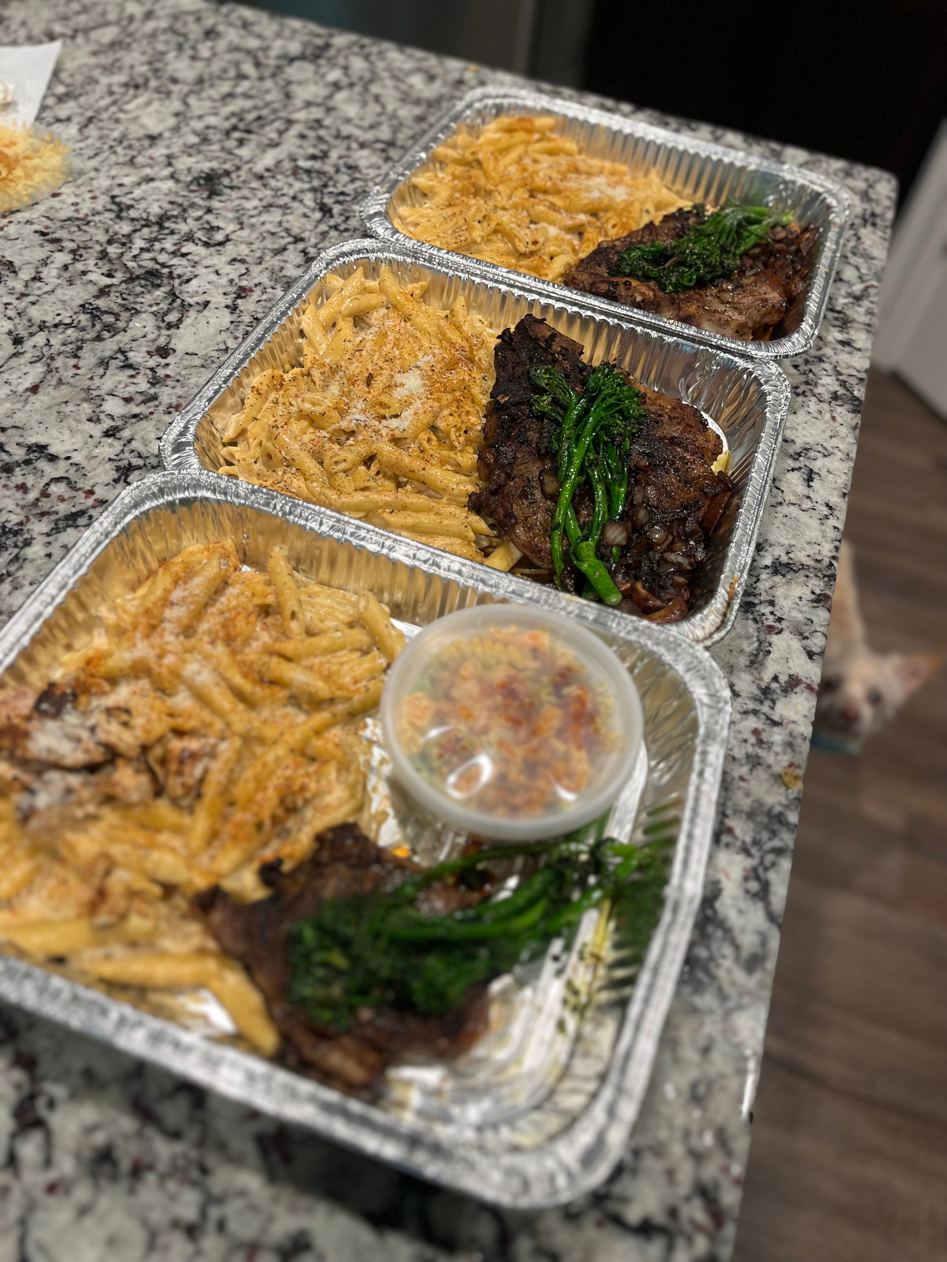 Three aluminum trays with cooked food on a granite countertop, including fried items, beef with greens, and pasta with sauce.