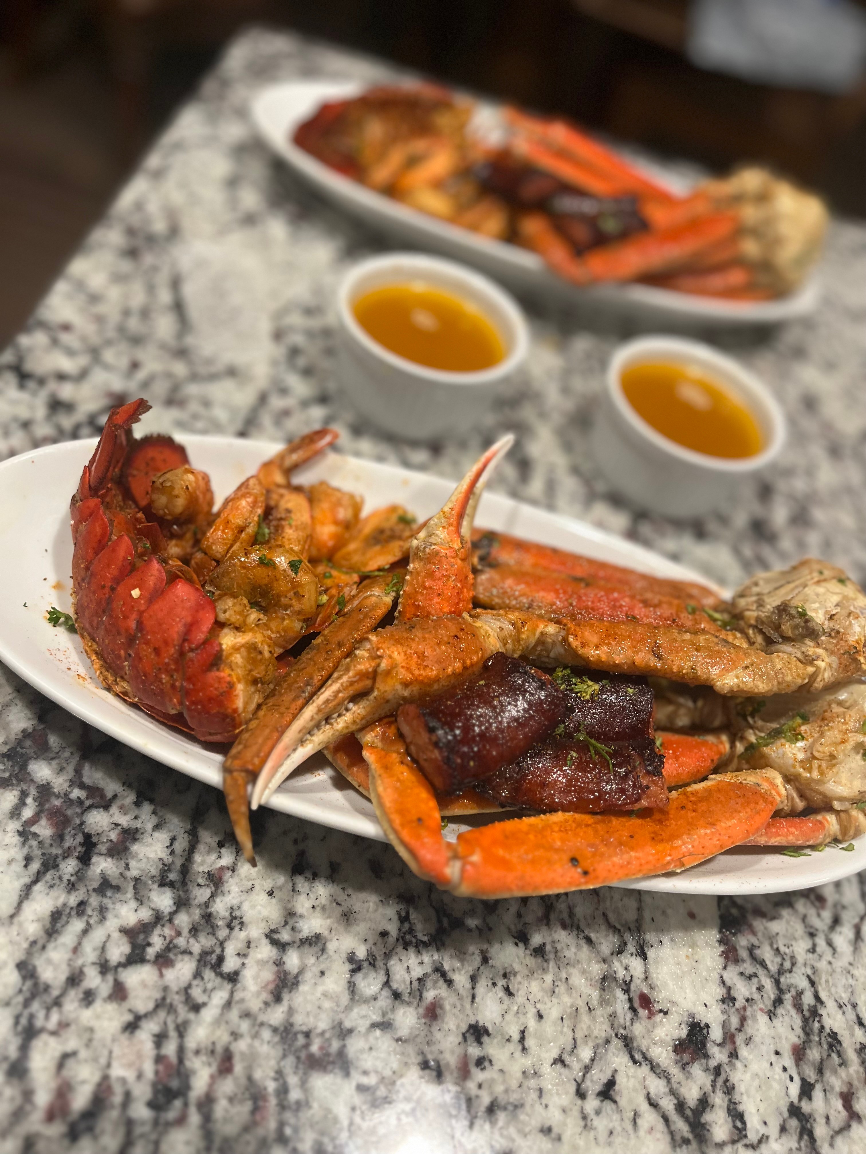 Seafood platter with lobster, crab, and shrimp on a white oval plate, two small bowls of sauce, on a gray marble table.