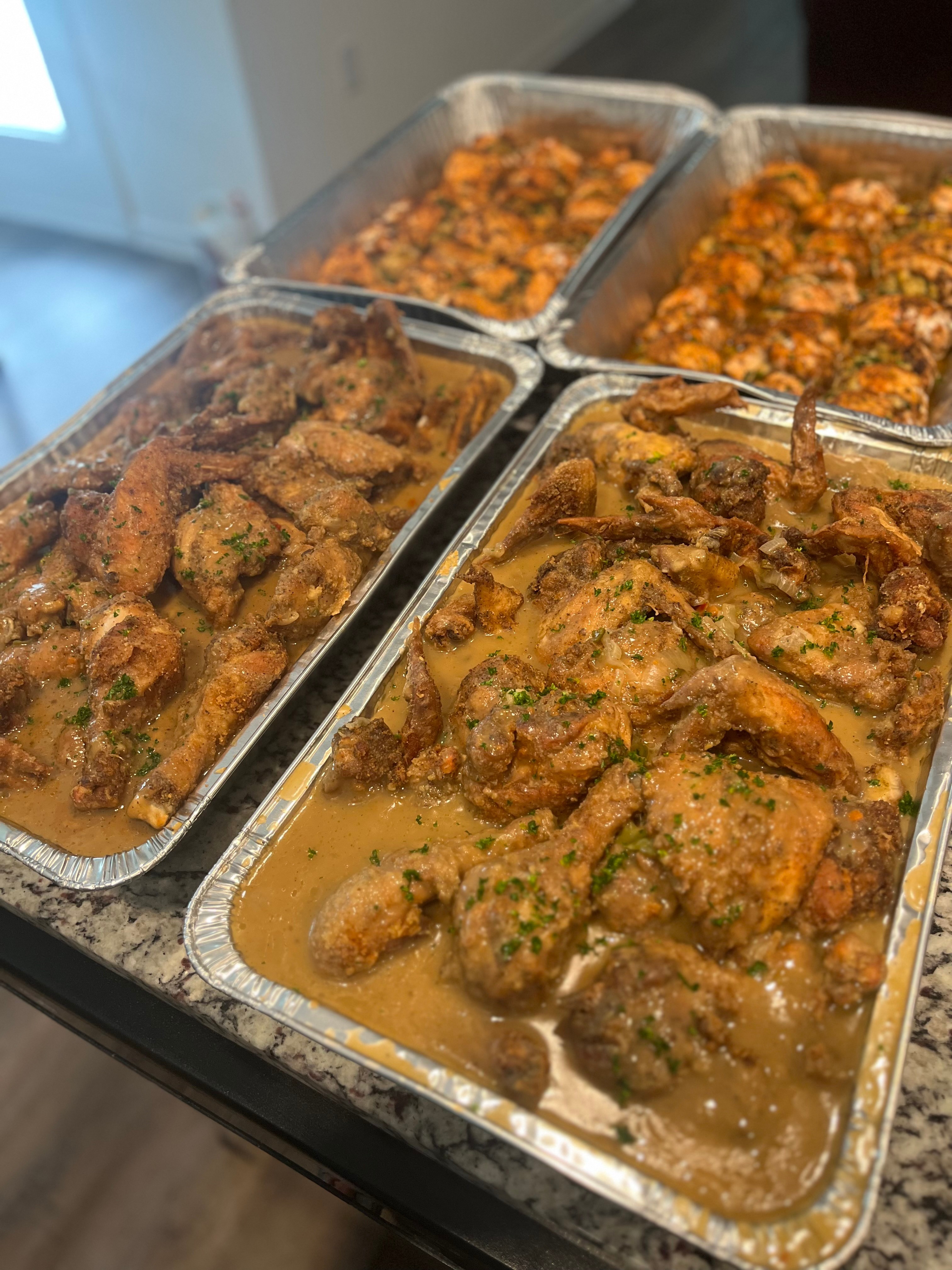 Four aluminum trays filled with cooked chicken dishes on a dark surface, with a blurred background of a room and window.