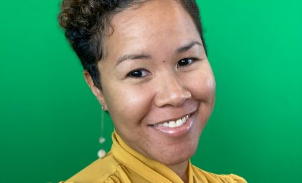 Smiling woman with short curly hair wearing a yellow blouse against a green background.