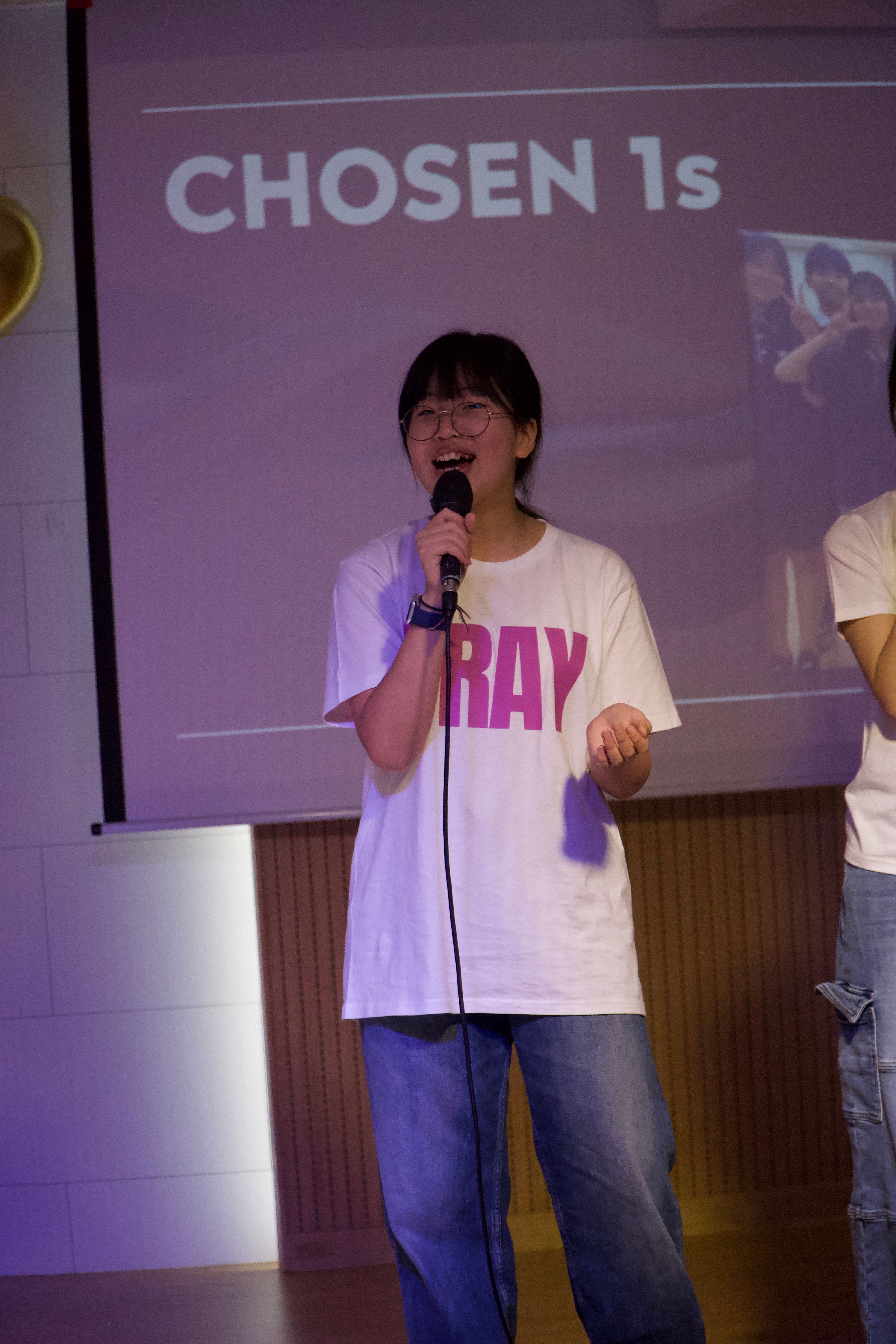 Person standing on stage with microphone, wearing white shirt and jeans, in front of a screen with text. Word count: 20