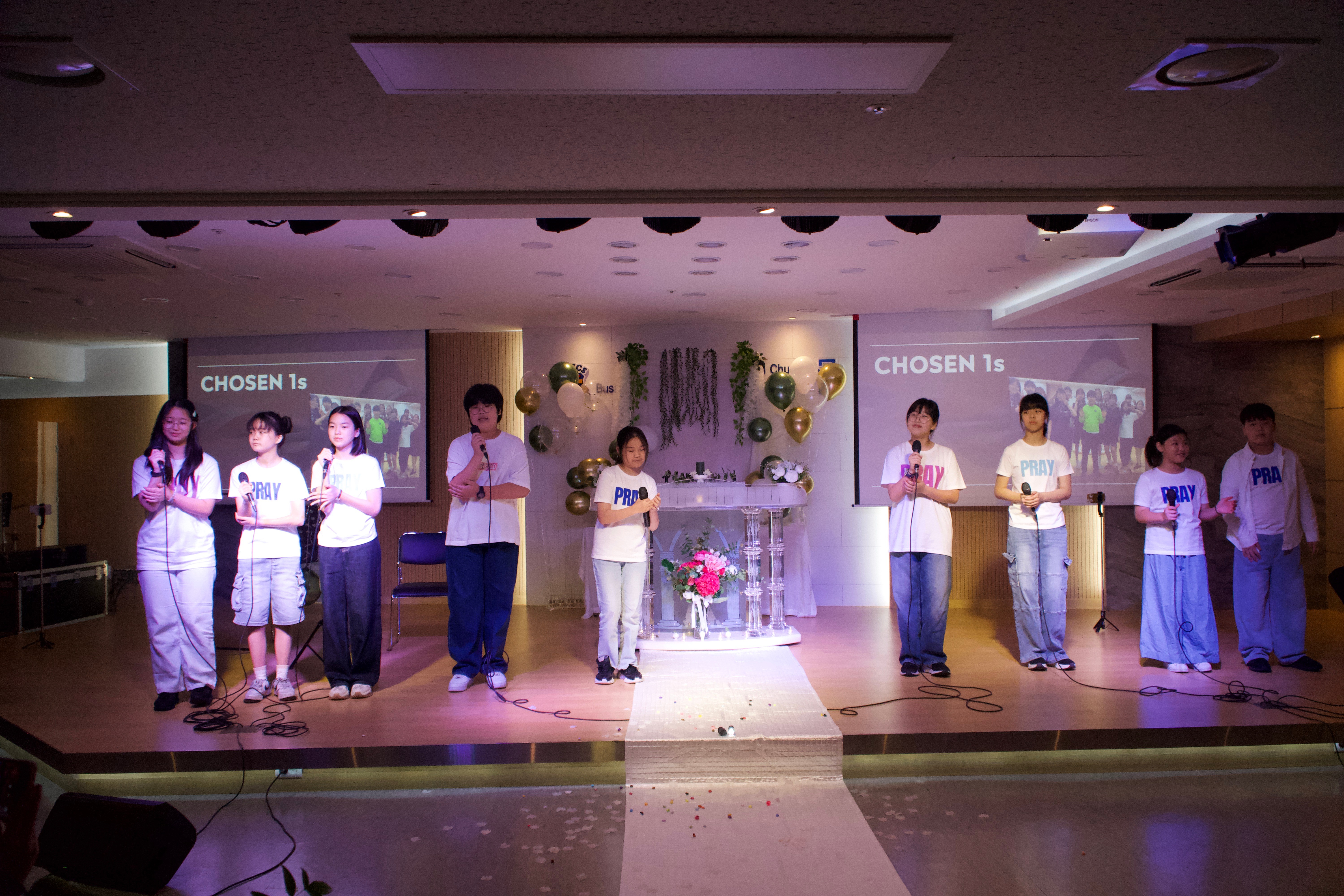 Group of children on stage holding papers, with a decorated backdrop and screens displaying text, in a well-lit room.