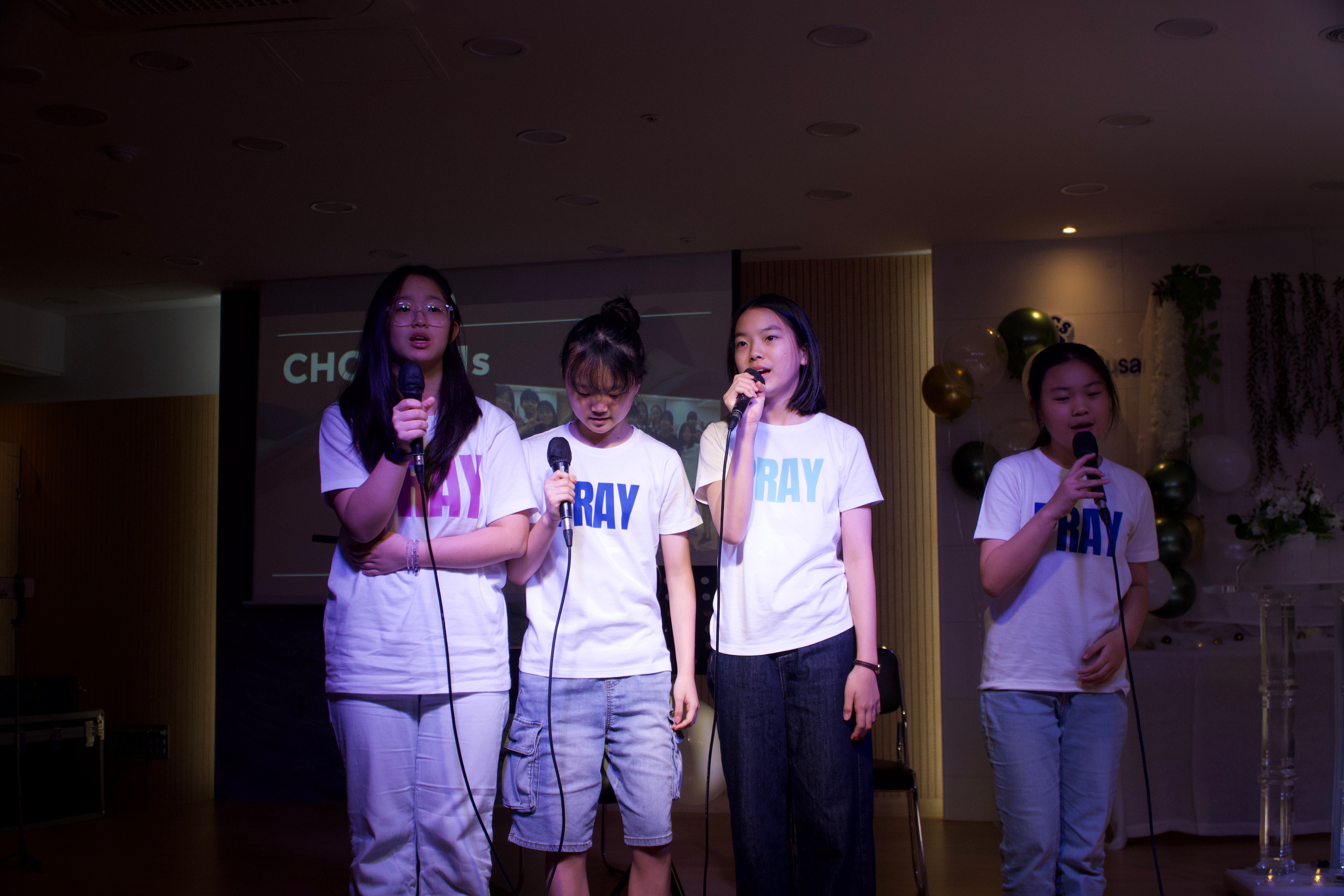 Four young girls stand on stage holding microphones, wearing white T-shirts with blue text, performing together.