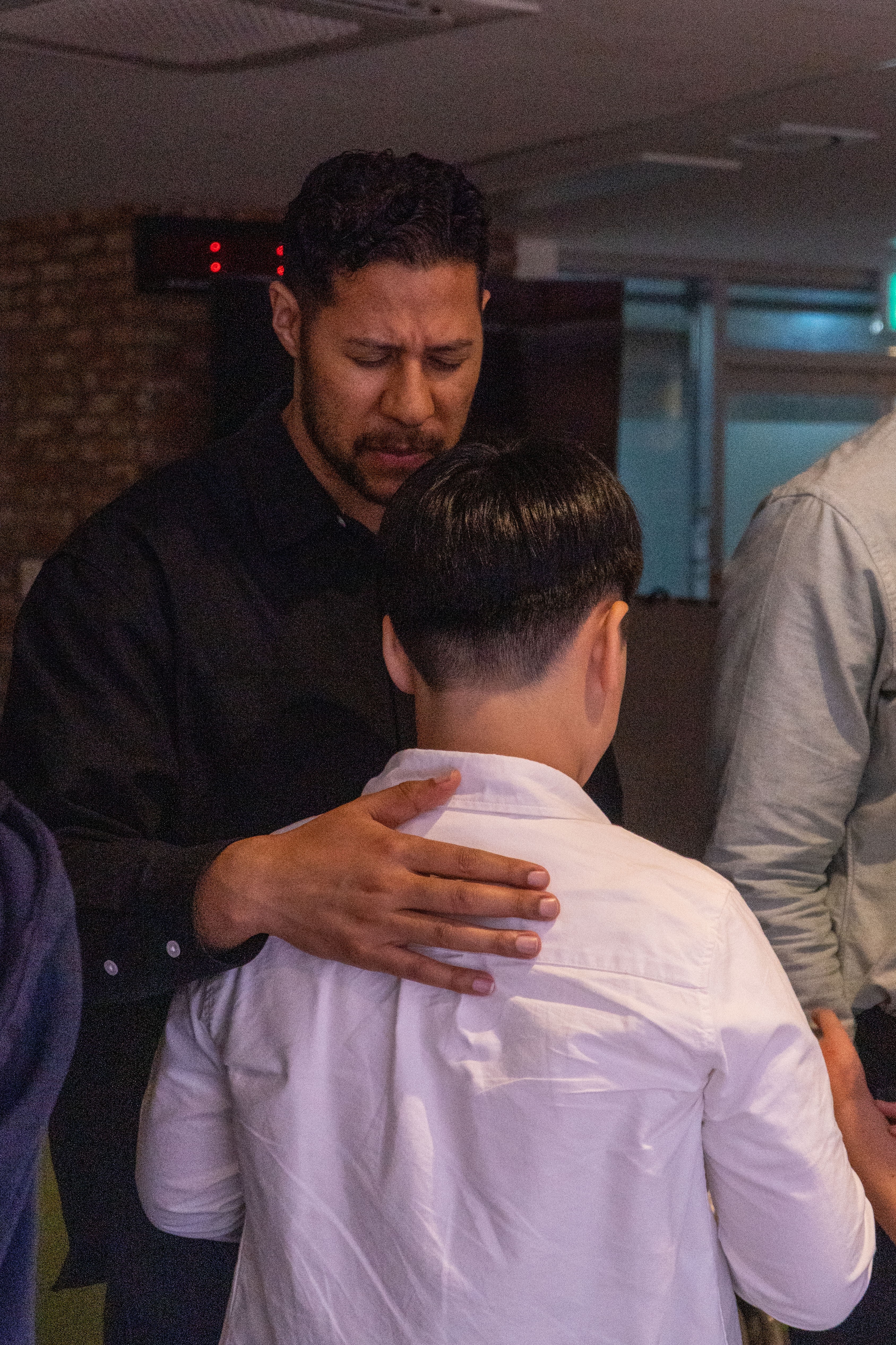 Man with dark hair and beard gently touches a person's shoulder in a white shirt indoors.