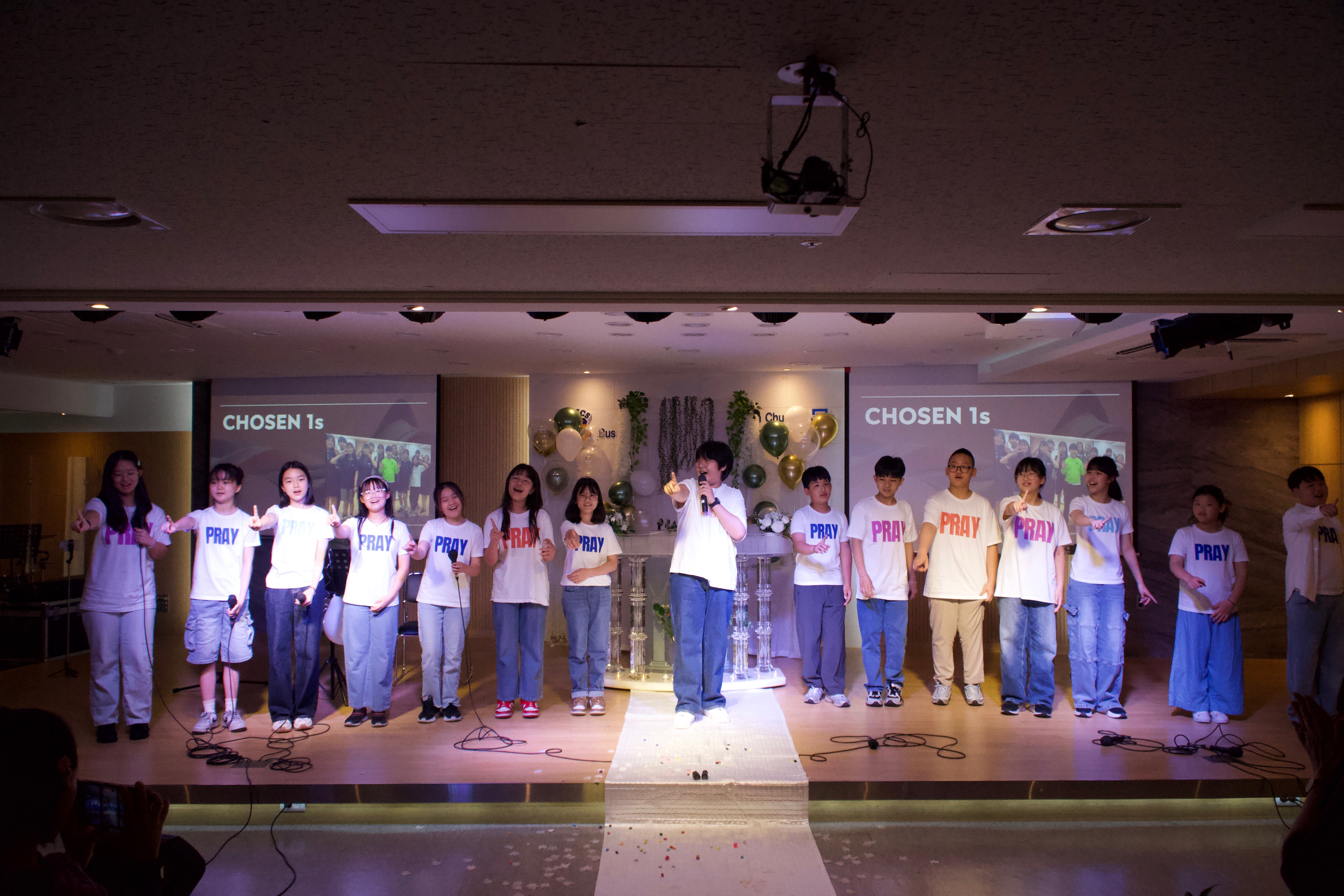 Group of children standing on stage, some holding microphones, with a backdrop and stage lighting, in a school performance.