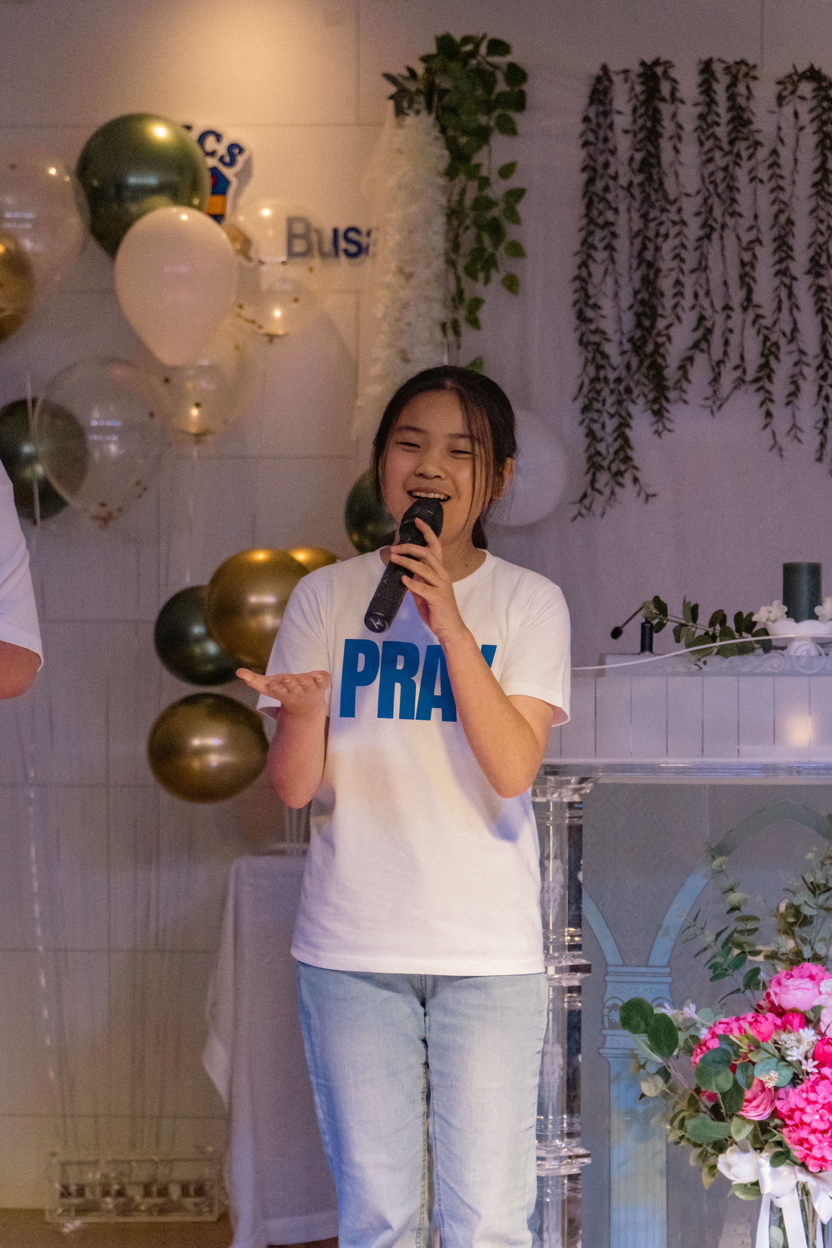Girl holding microphone and smiling at celebration with balloons and flowers in background.
