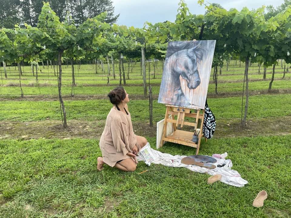 Person kneeling on grass in vineyard, painting horse on canvas, with art supplies nearby, trees in background.