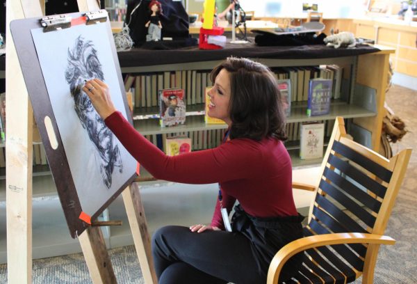 Woman drawing a lion on an easel in a room with shelves and art supplies, sitting on a chair.