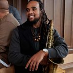 Person with dreadlocks holding a saxophone, smiling, wearing a black jacket and watch, in a wooden-paneled room.