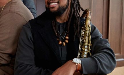 Person with dreadlocks holding a saxophone, smiling, wearing a black jacket and watch, in a wooden-paneled room.