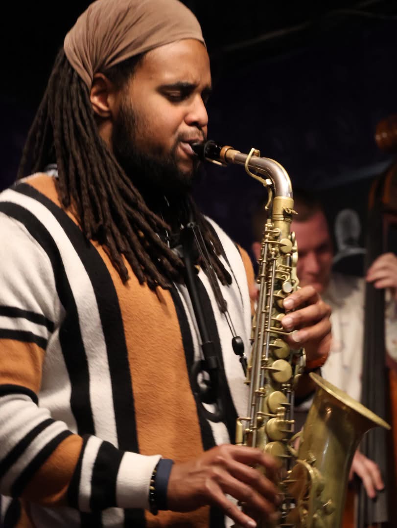 Person playing saxophone, wearing a headscarf and striped shirt, in a dimly lit setting.