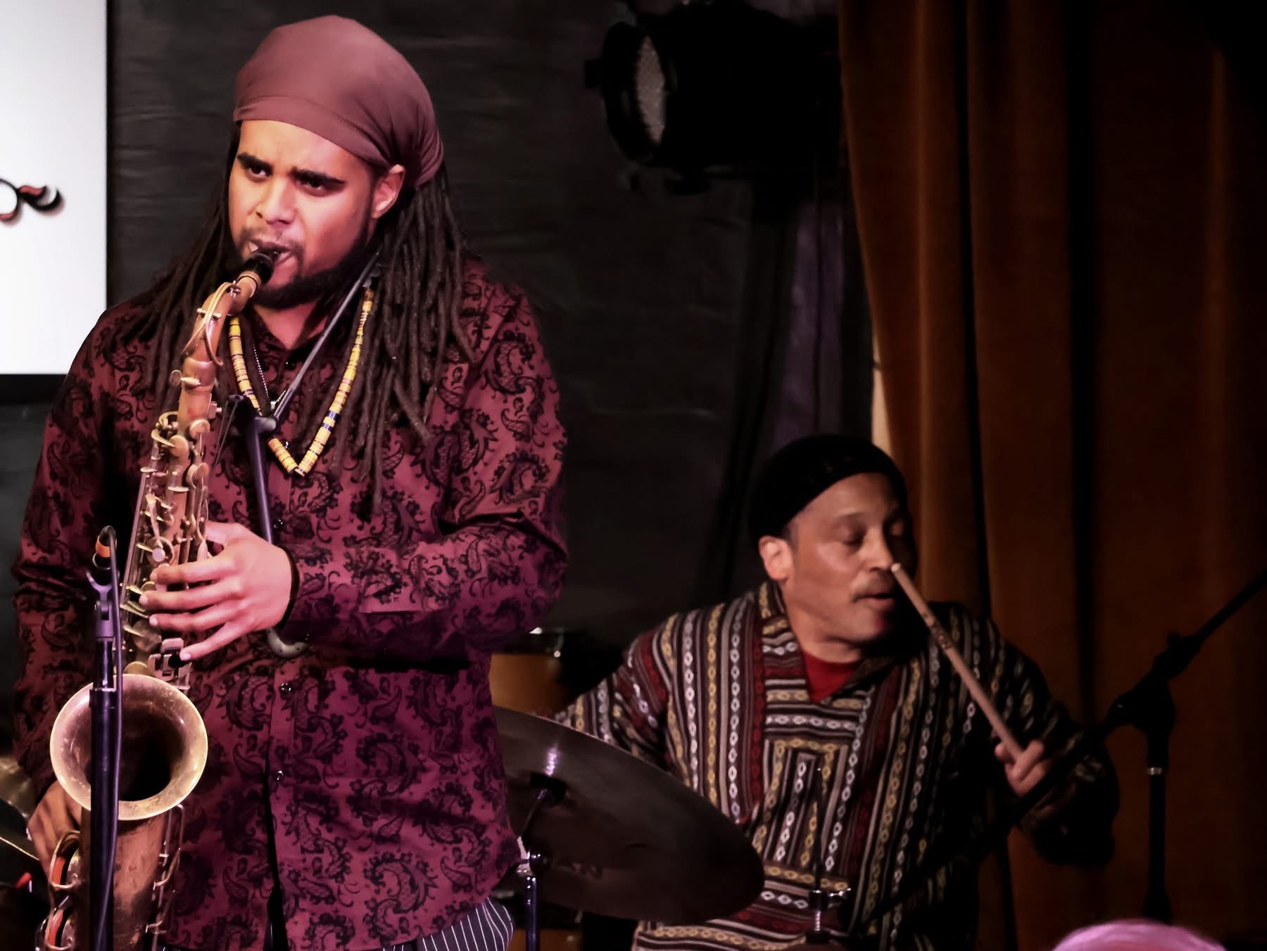Two musicians perform on stage, one playing a saxophone and the other a drum, with dark background and curtains.