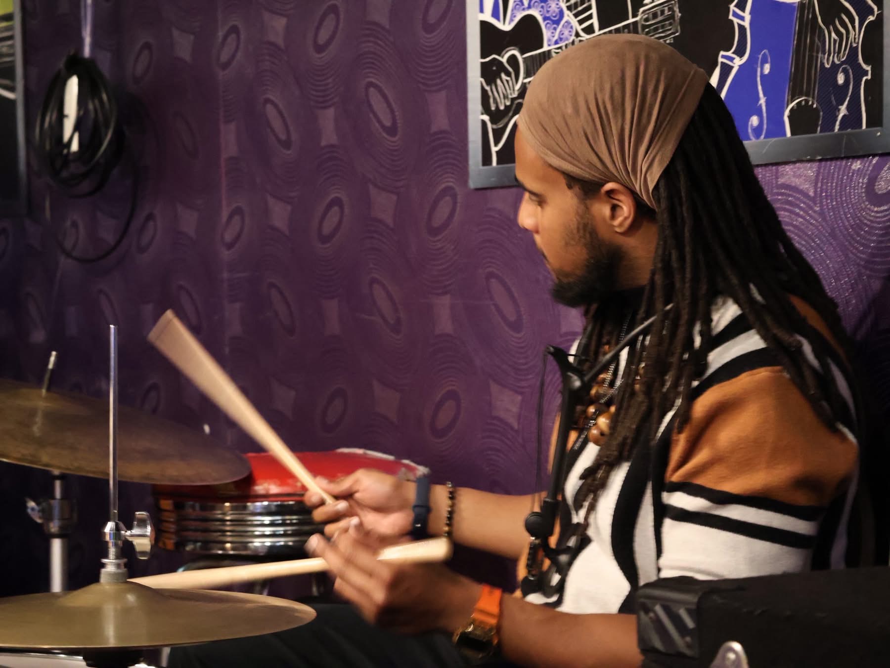 Person with long dreadlocks playing drums in a purple room with artwork on the wall.