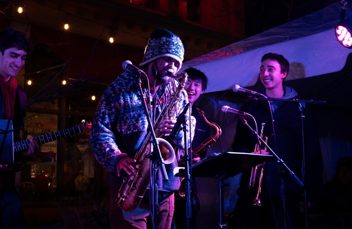Musician with a hat playing saxophone on stage with three smiling band members, colorful lighting, and a dark background.
