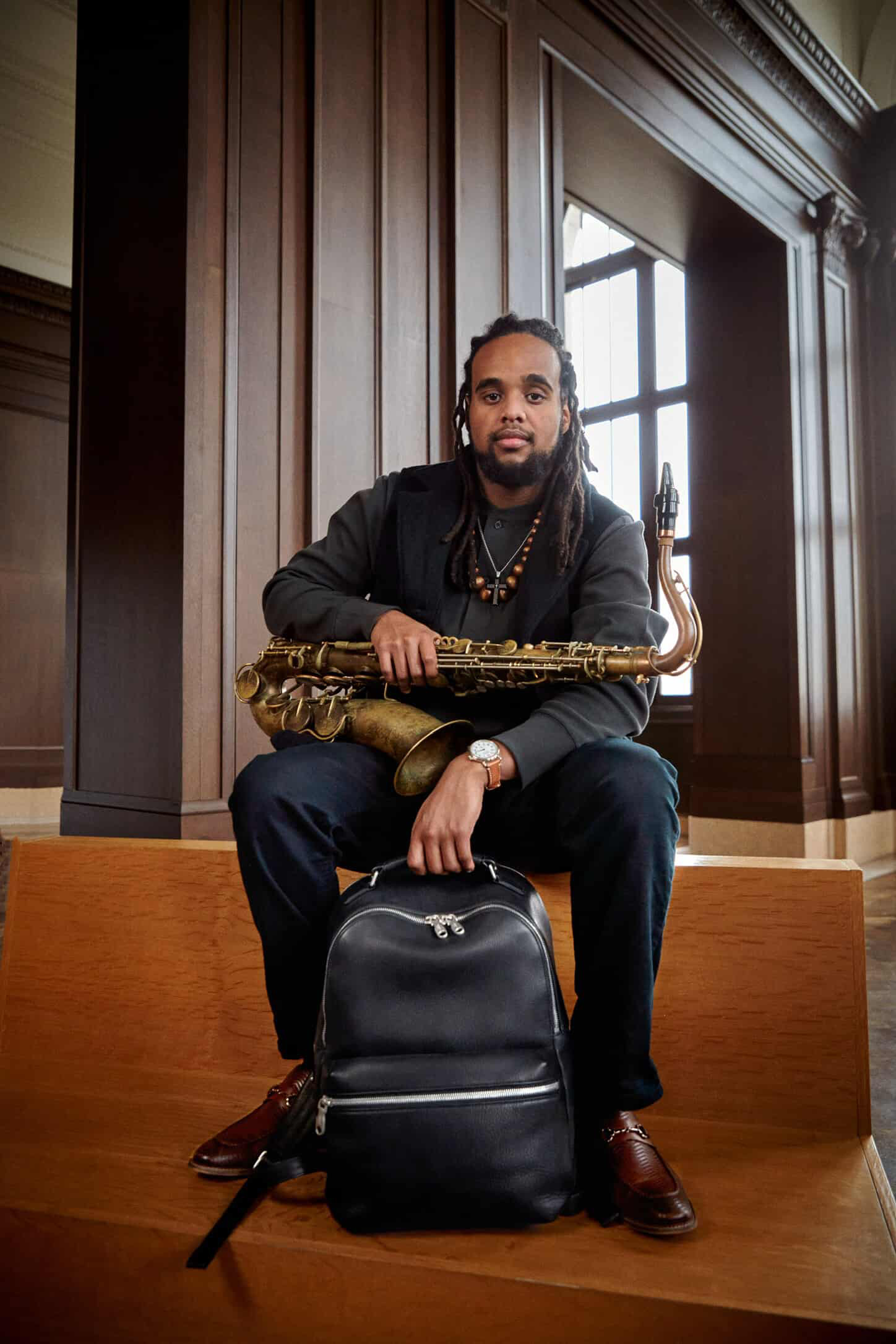 Man with dreadlocks and beard sitting on a wooden bench holding a saxophone, with a black backpack in front, in a room with wooden paneling and large window.