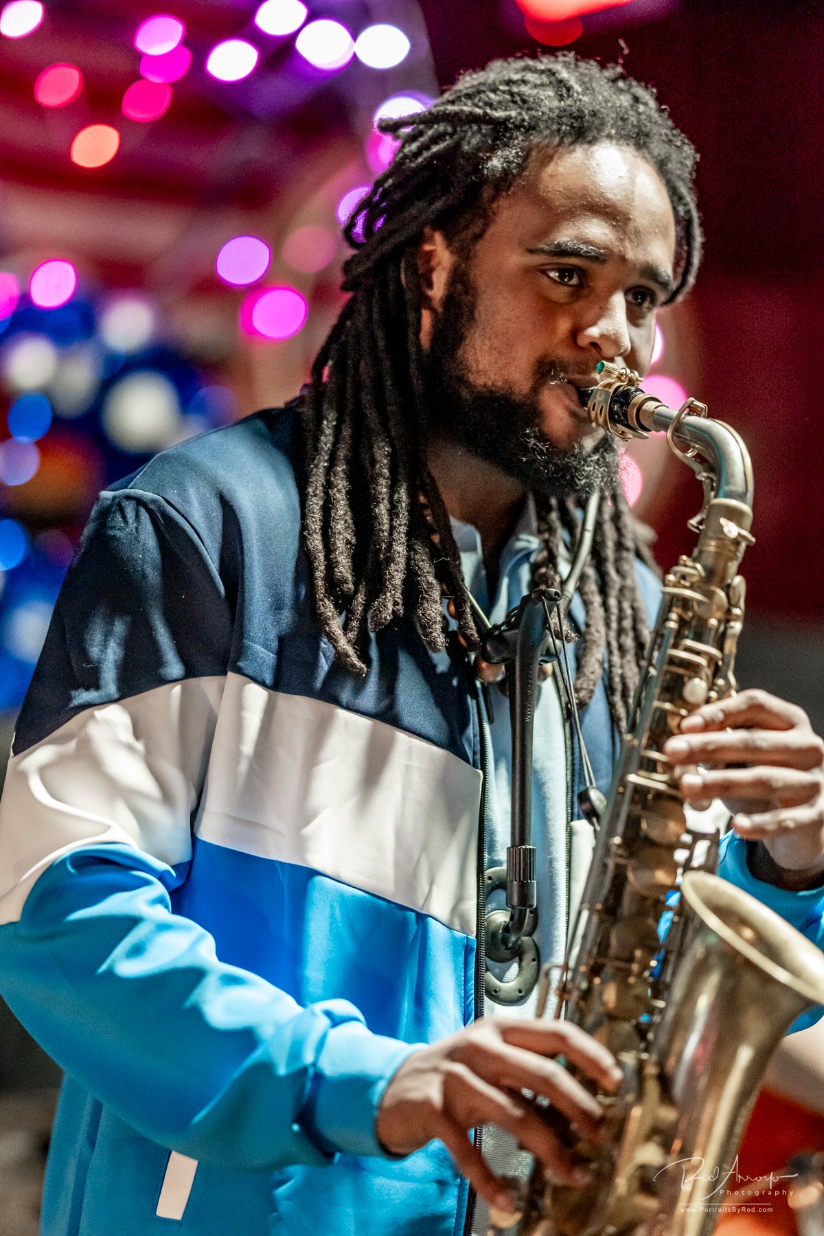 Person with dreadlocks playing a saxophone on stage with colorful lights in background.
