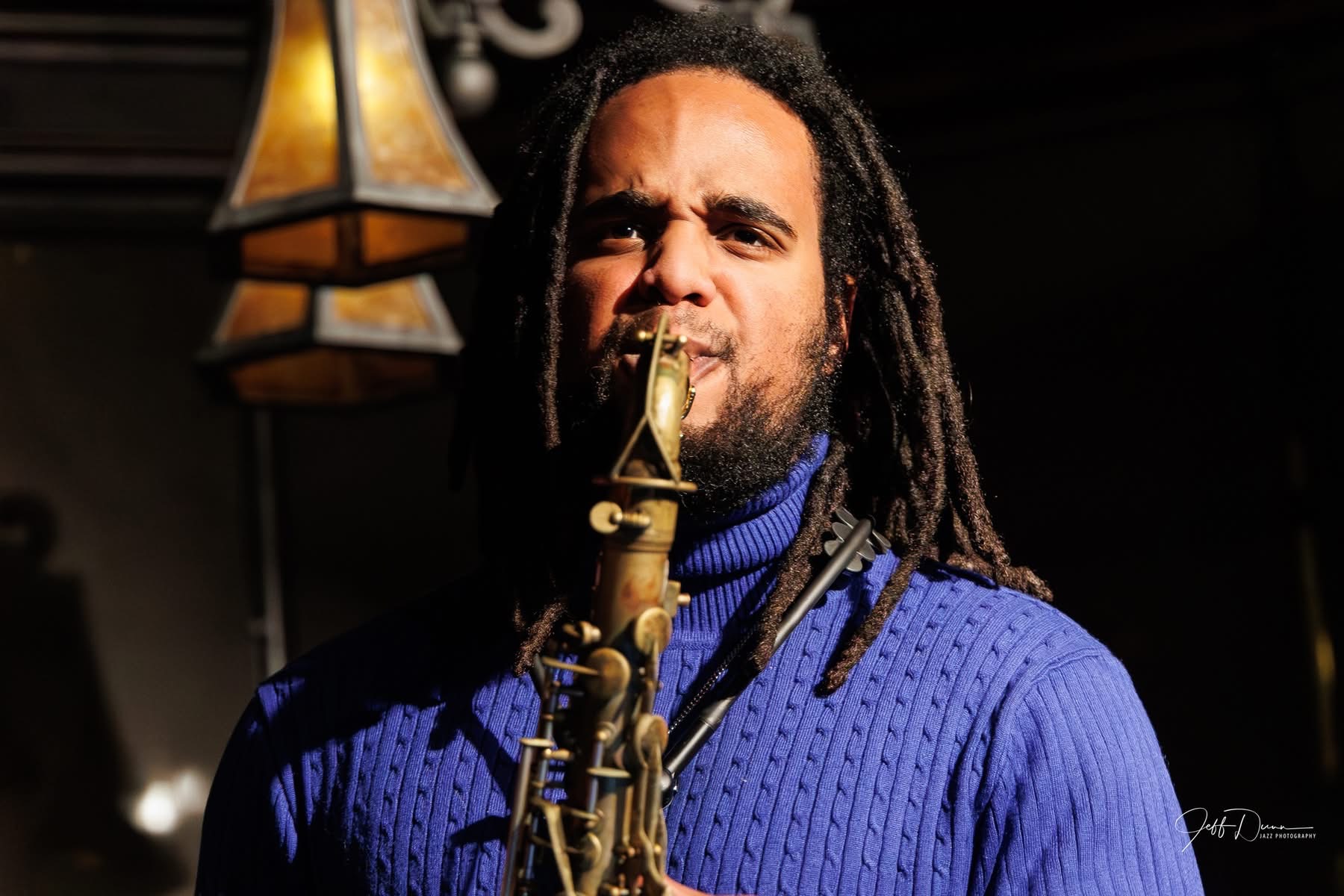 Person with dreadlocks playing a saxophone, wearing a blue sweater, in a dimly lit setting.