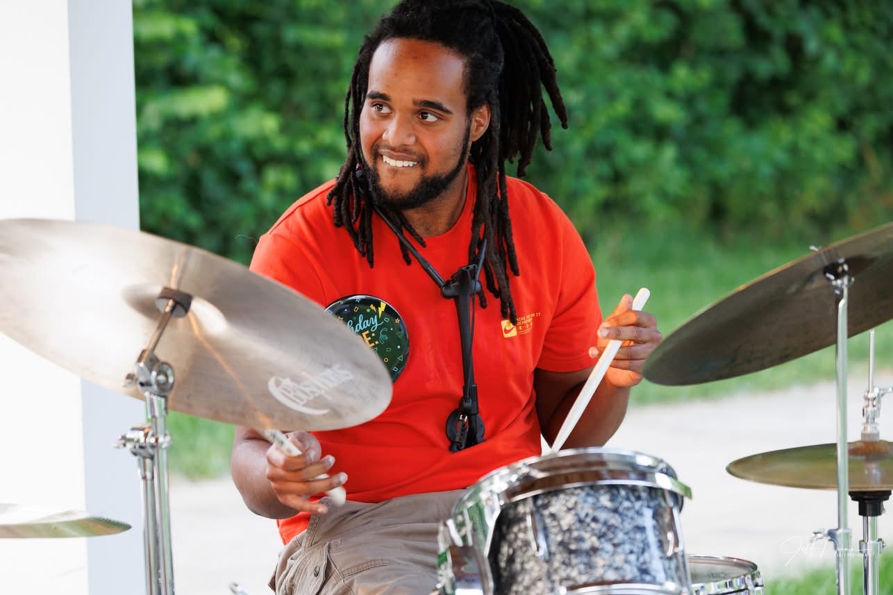 Person with dreadlocks playing drums outdoors, smiling, wearing a red shirt, with green trees in background.