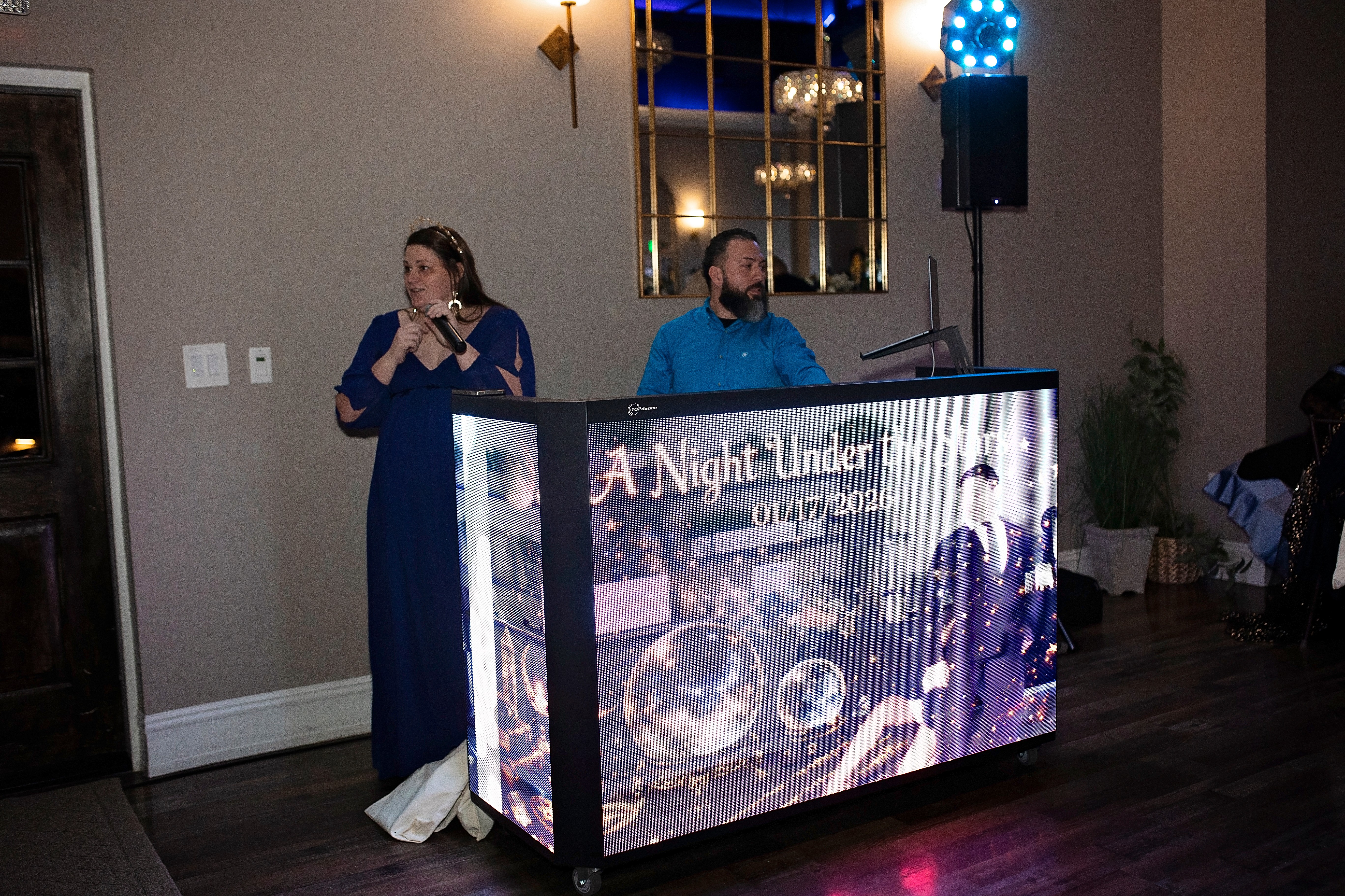 A woman and a man stand behind a DJ booth with a colorful display, in a decorated indoor event space.