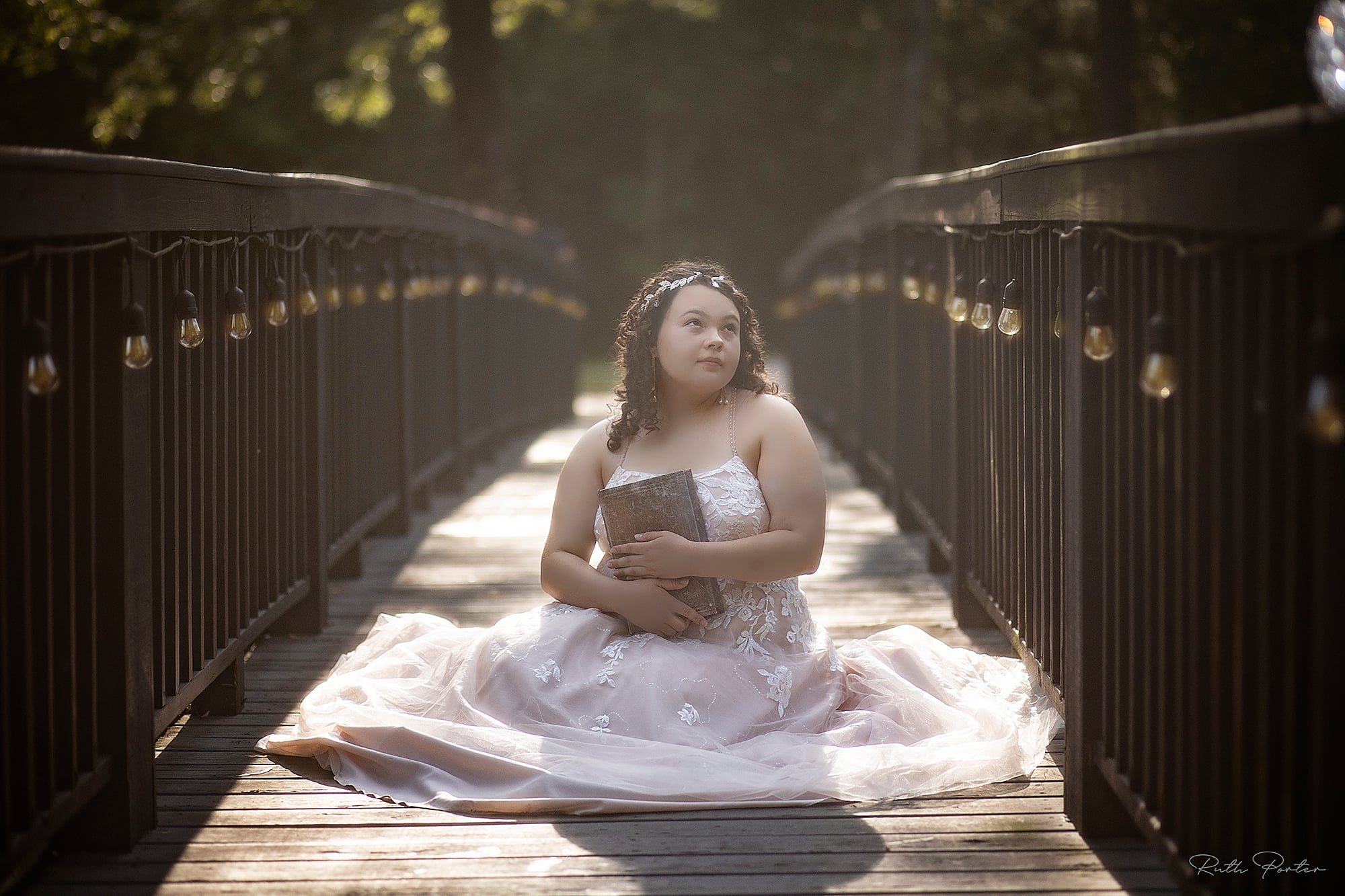 Young girl in a white dress sitting on a bridge with railings and string lights, holding her hands together, looking to the side.