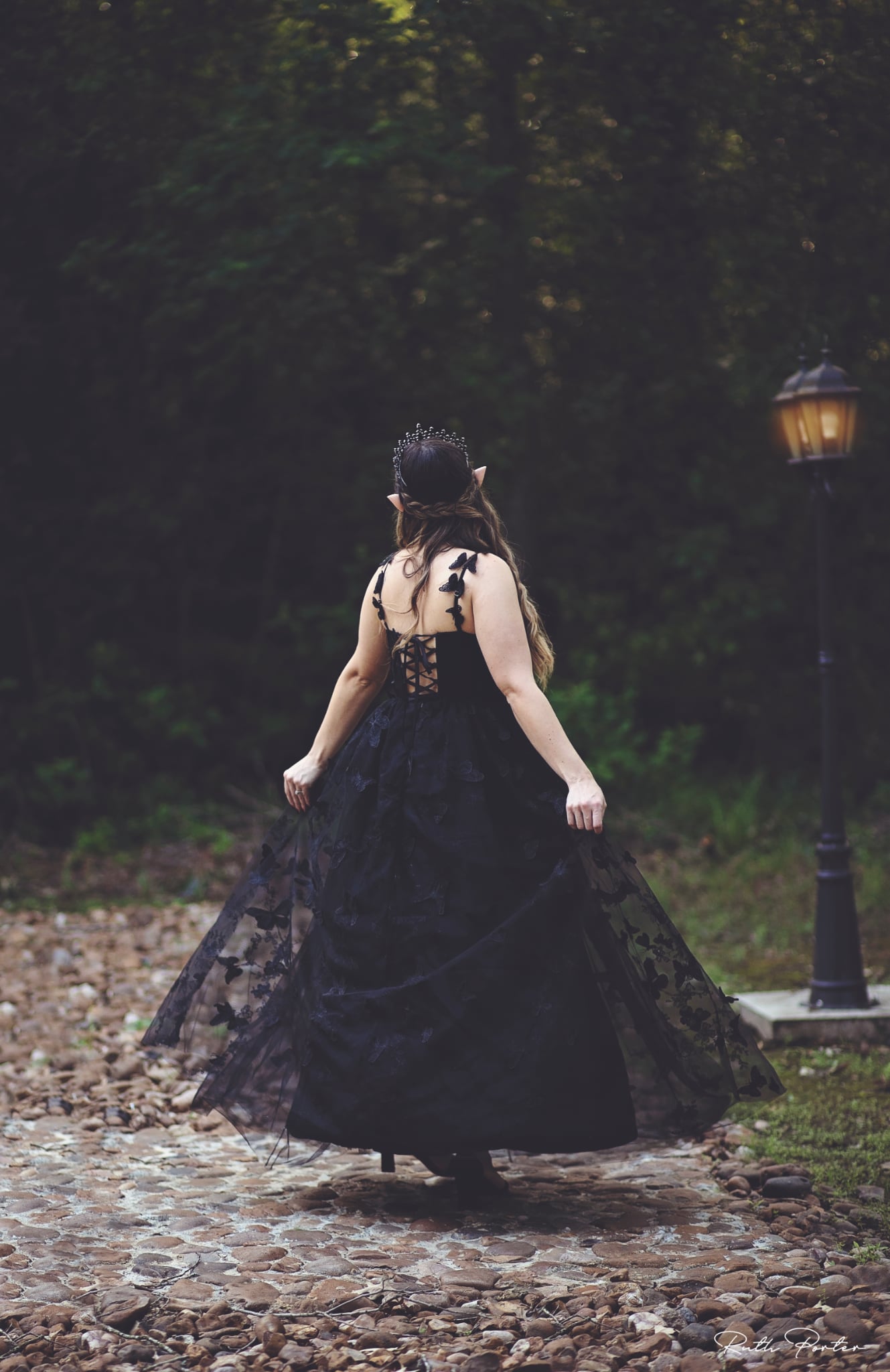 Person in a black dress walking on a cobblestone path with trees and a lamp post in the background.