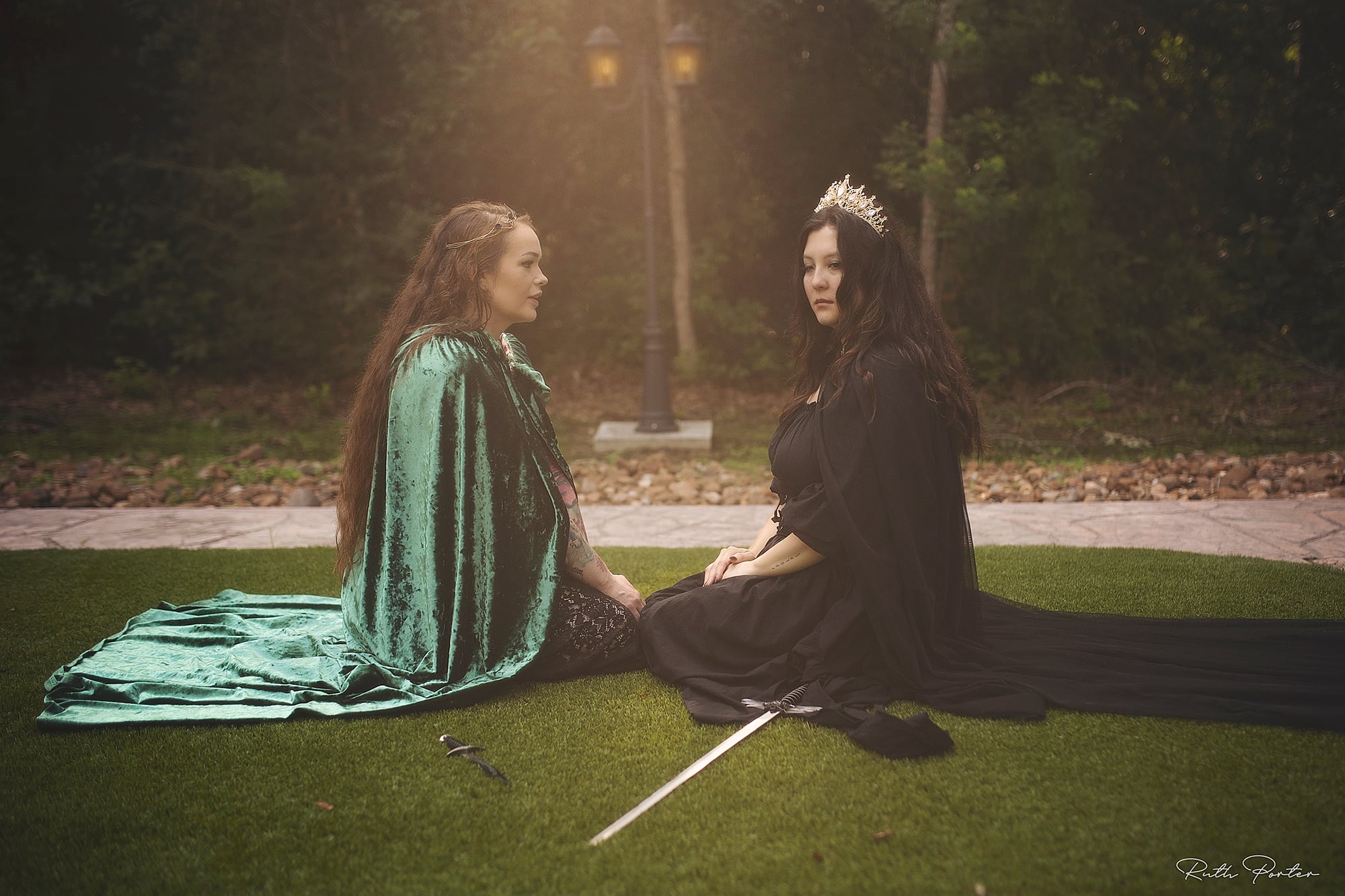 Two women sit on grass, one wearing a crown, in a park with trees and a bench in the background.