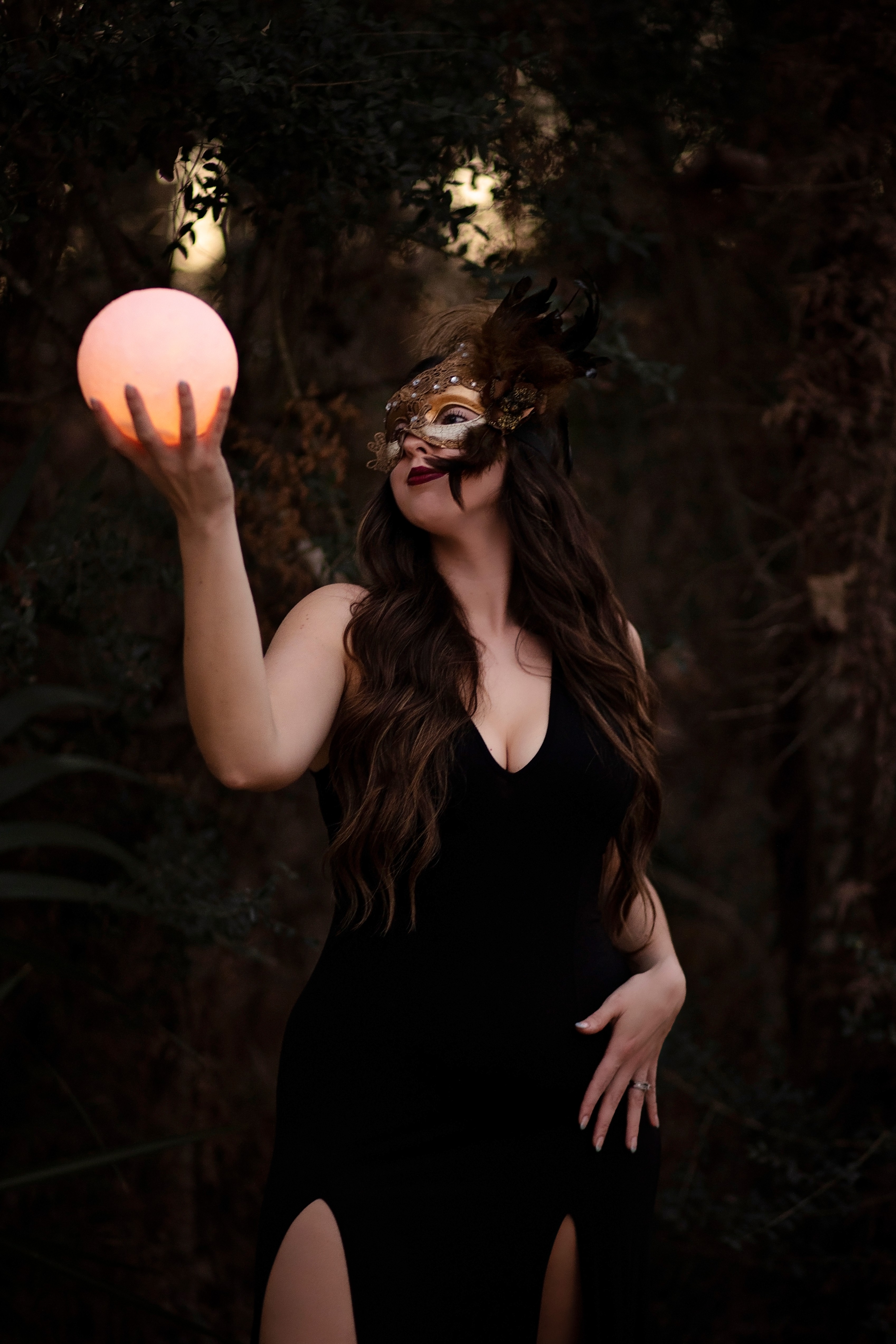Woman in black dress holding glowing pink orb outdoors at night, wearing a mask, with dark trees in background.