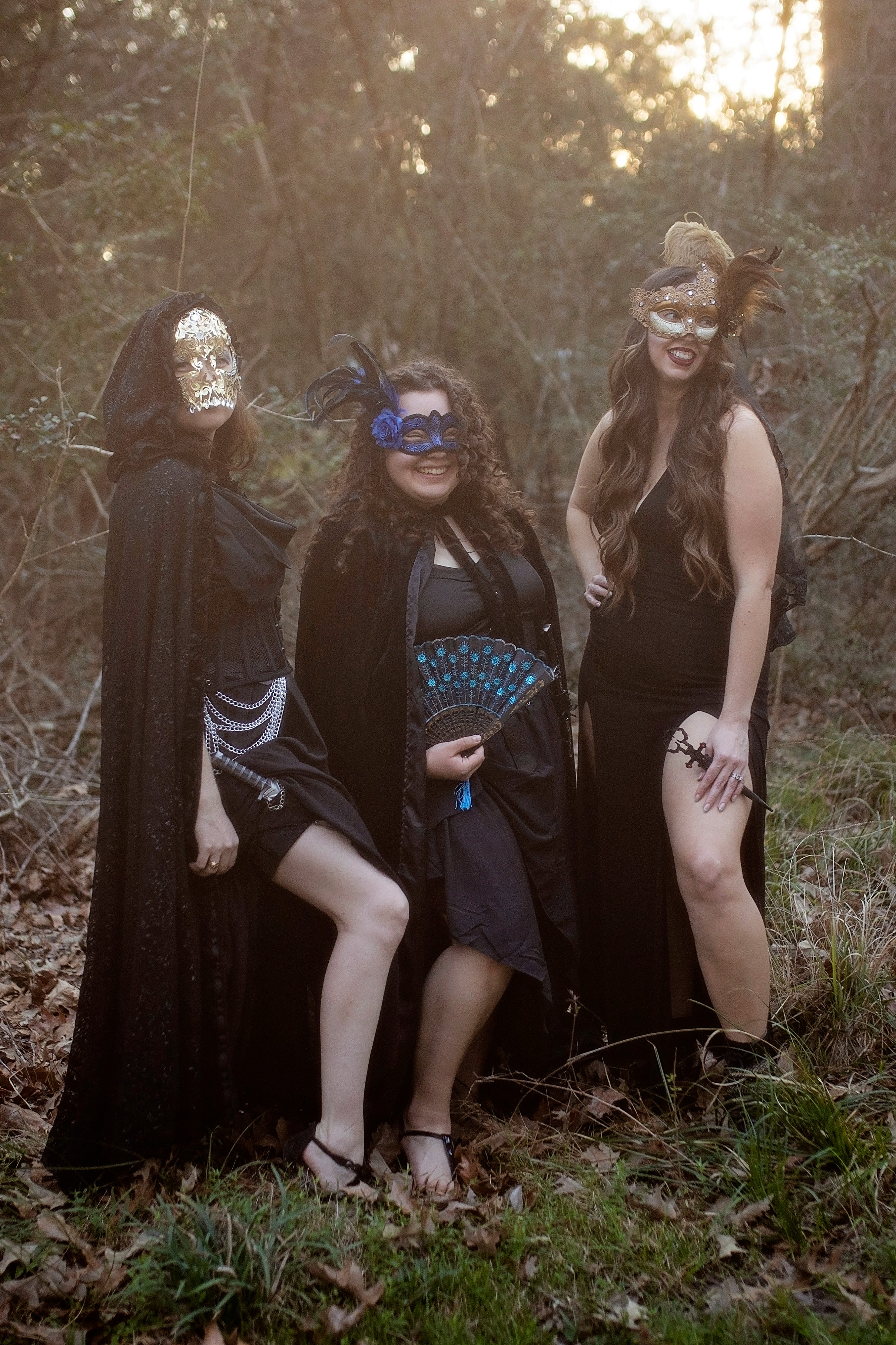 Three people in costumes with masks, standing outdoors in a wooded area, dressed in dark clothing and accessories.