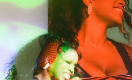 Two women singing into microphones, one smiling with eyes closed, wearing a mesh top and Harley-Davidson shirt.