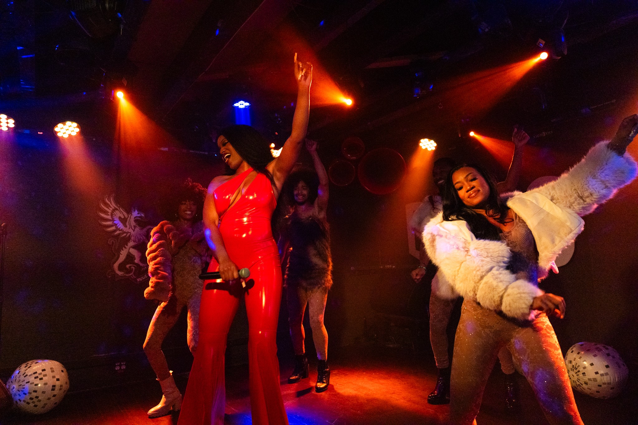 Group of women dancing on stage with colorful lights, some wearing bright outfits and fur coat, lively atmosphere.