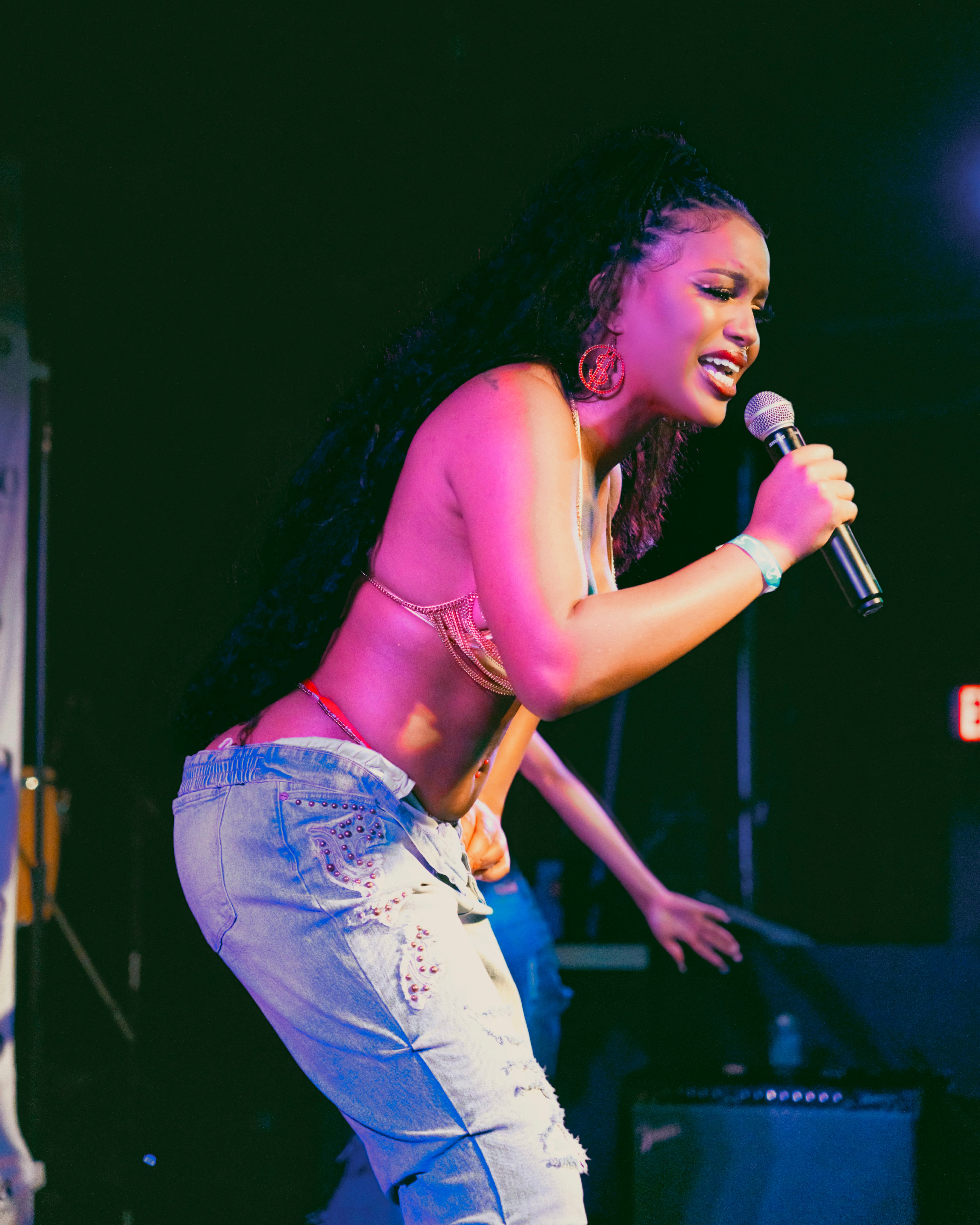 Performer singing into a microphone on stage, wearing a pink top and ripped jeans, with a dark background.