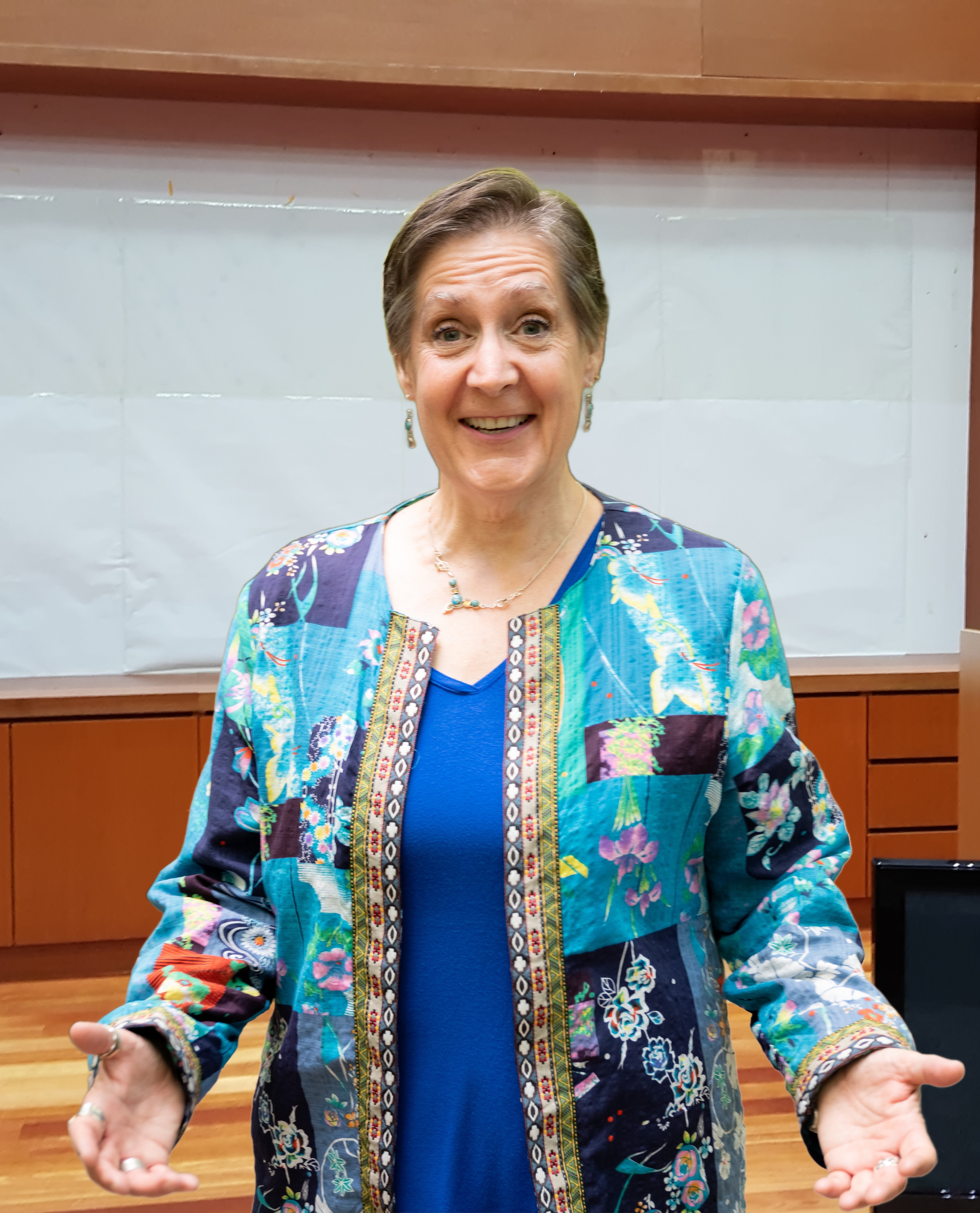 Smiling woman with short hair wearing a colorful patterned jacket and blue top, standing indoors in front of a whiteboard.