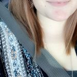 Close-up of a smiling young woman with light skin, brown hair, and a nose piercing, wearing a patterned top and seatbelt.