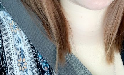 Close-up of a smiling young woman with light skin, brown hair, and a nose piercing, wearing a patterned top and seatbelt.