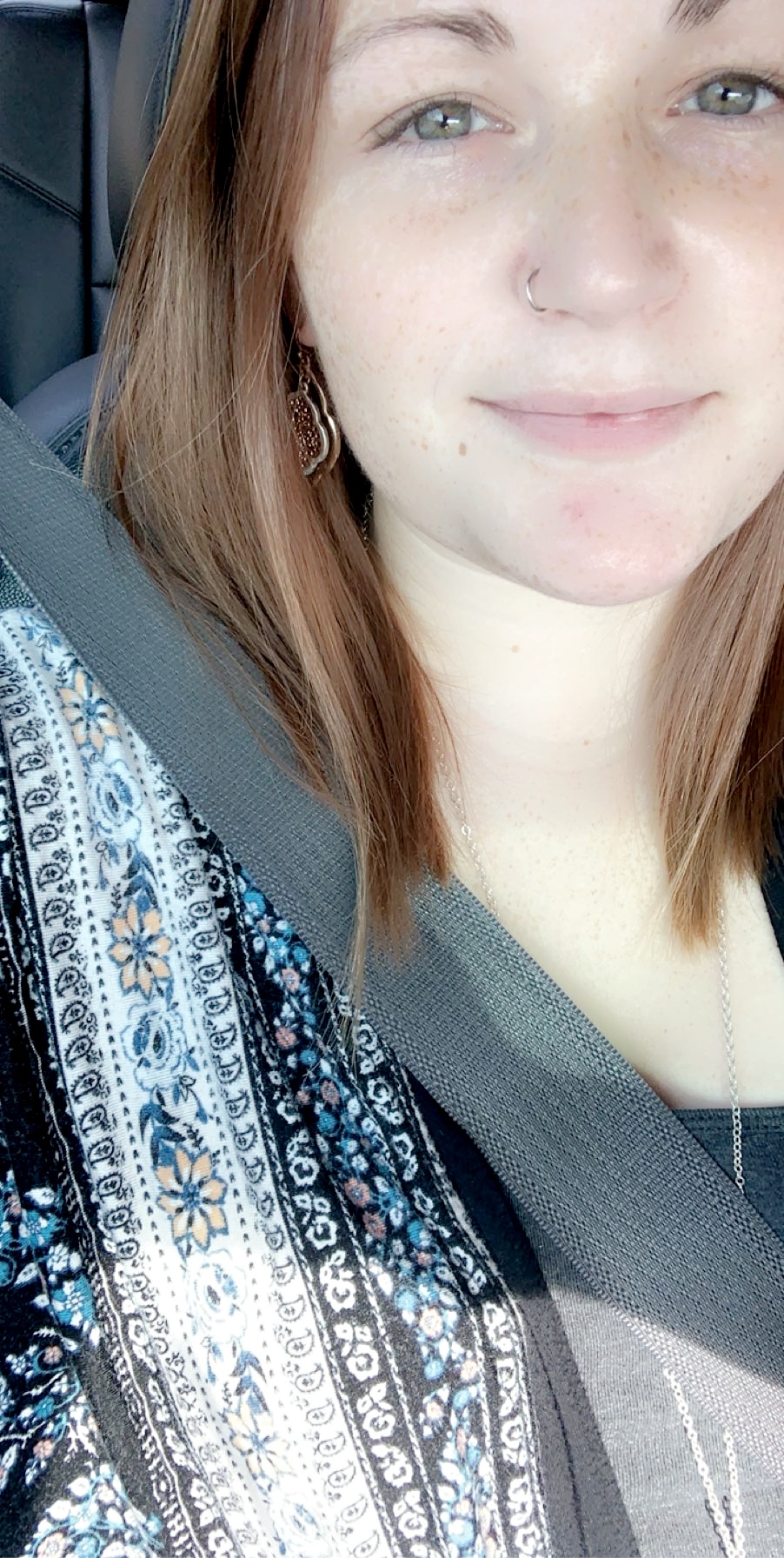 Close-up of a smiling young woman with light skin, brown hair, and a nose piercing, wearing a patterned top and seatbelt.