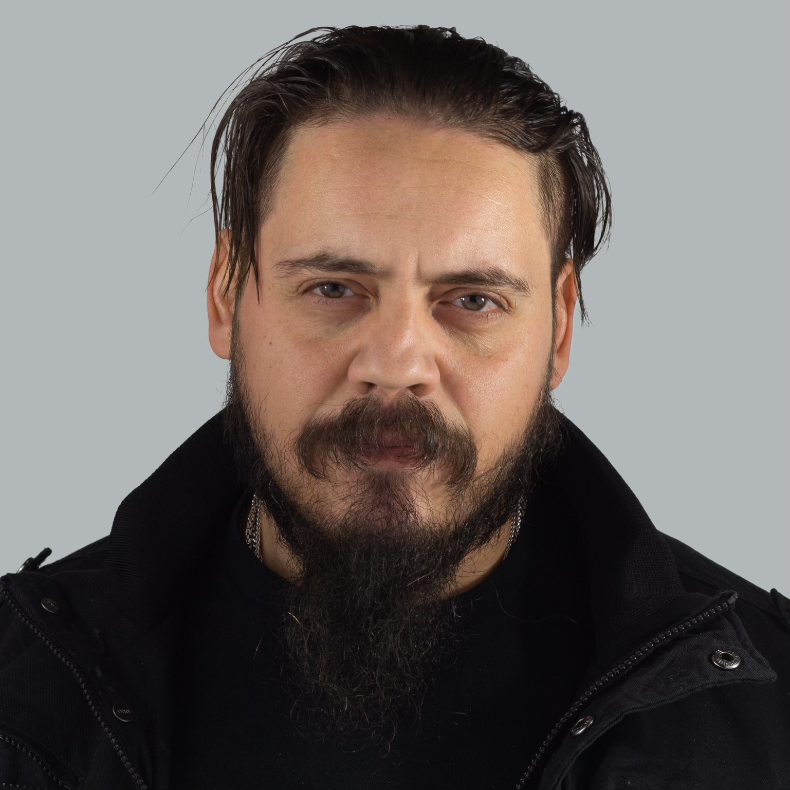 Man with dark hair, beard, and mustache wearing a black jacket against a gray background.