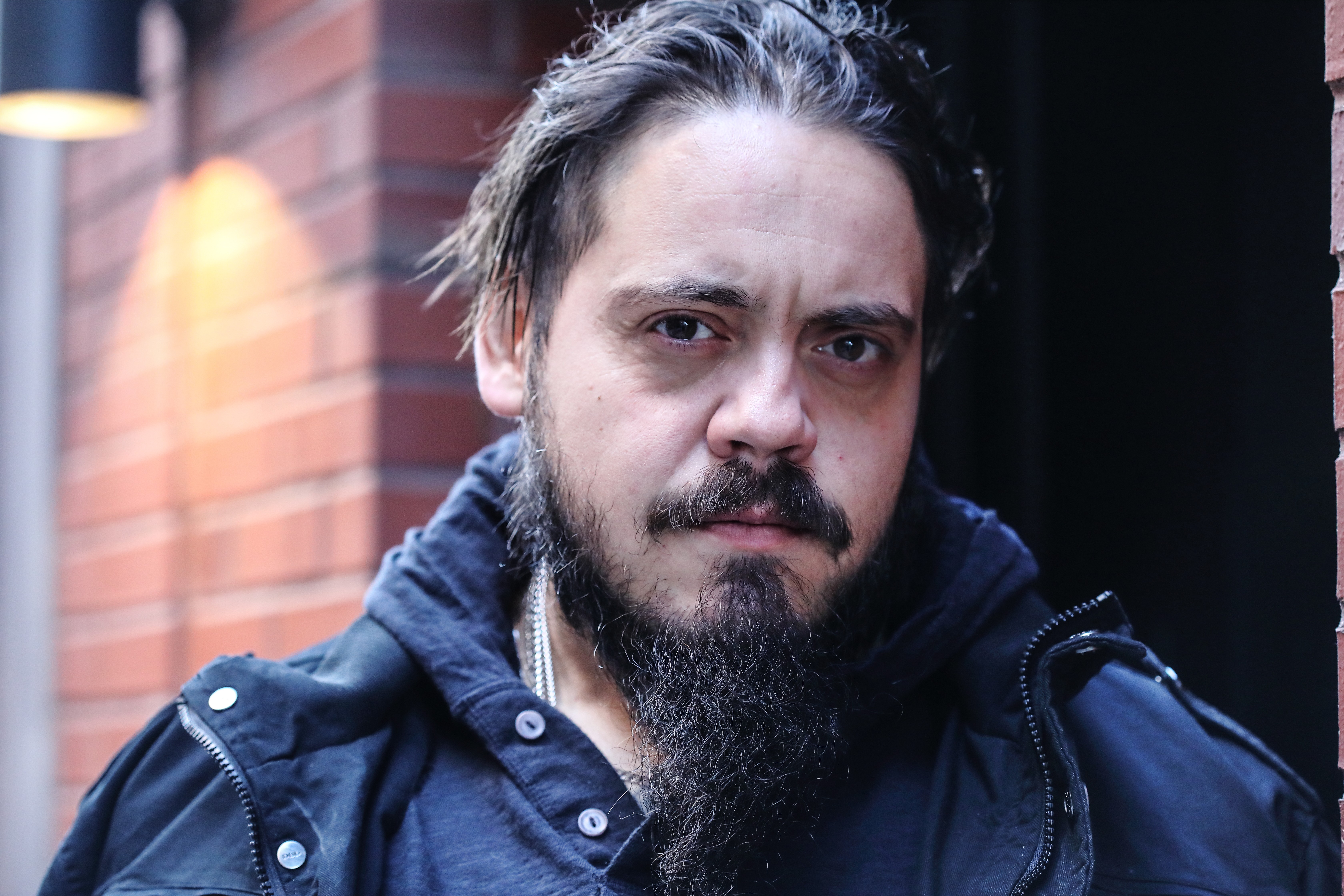 Man with dark hair and beard wearing a dark jacket, standing outdoors near a brick wall, looking at the camera.
