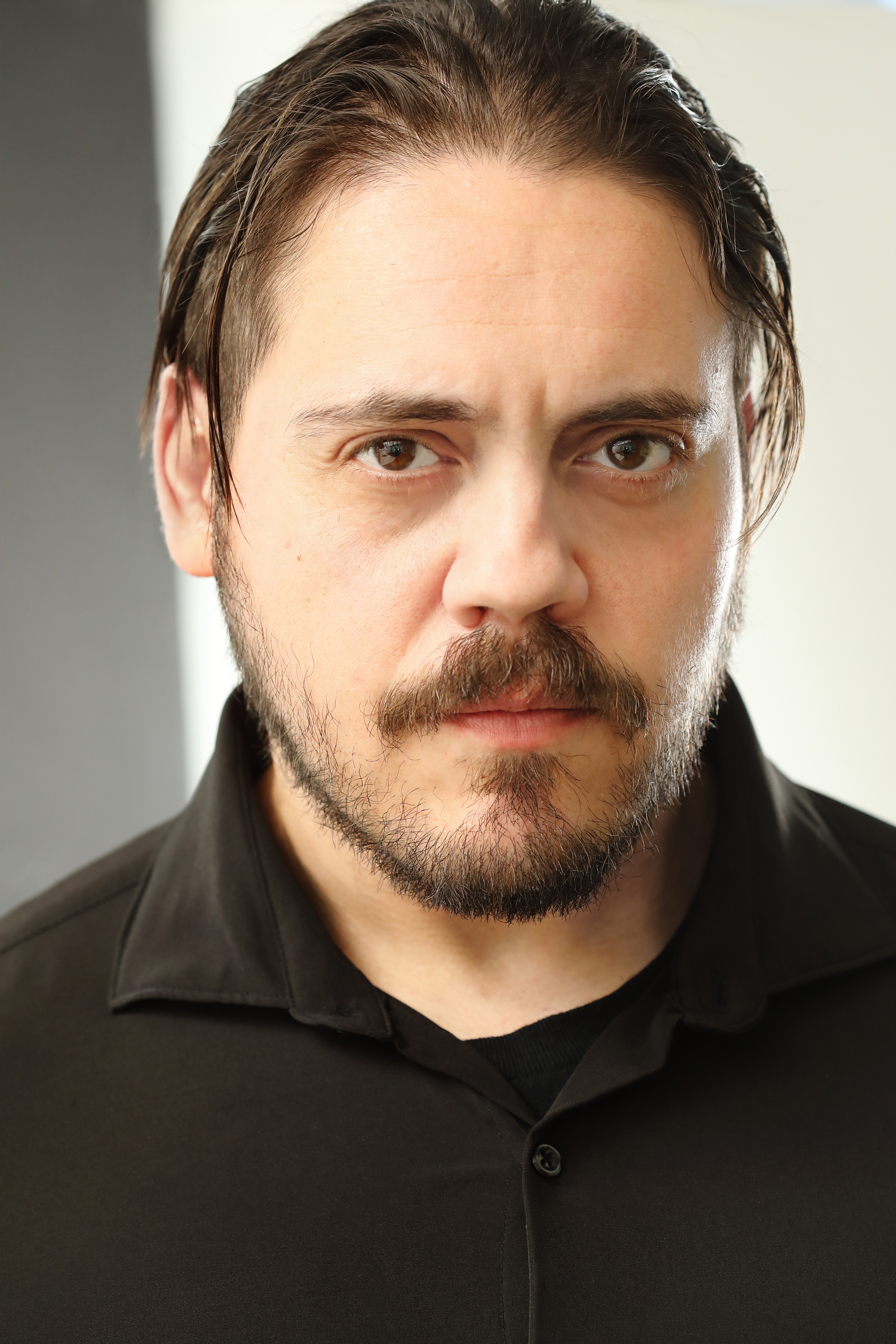 Close-up of a man with dark hair, beard, and mustache, wearing a black shirt, looking serious.