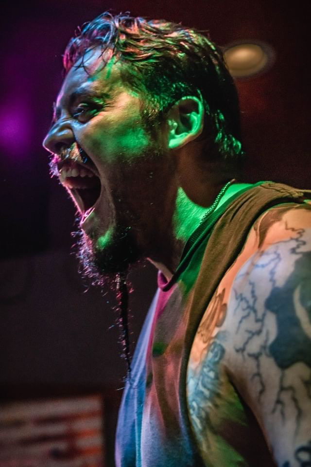 Man with a beard and tattoos, shouting or singing passionately, with colorful stage lighting on his face and hair.