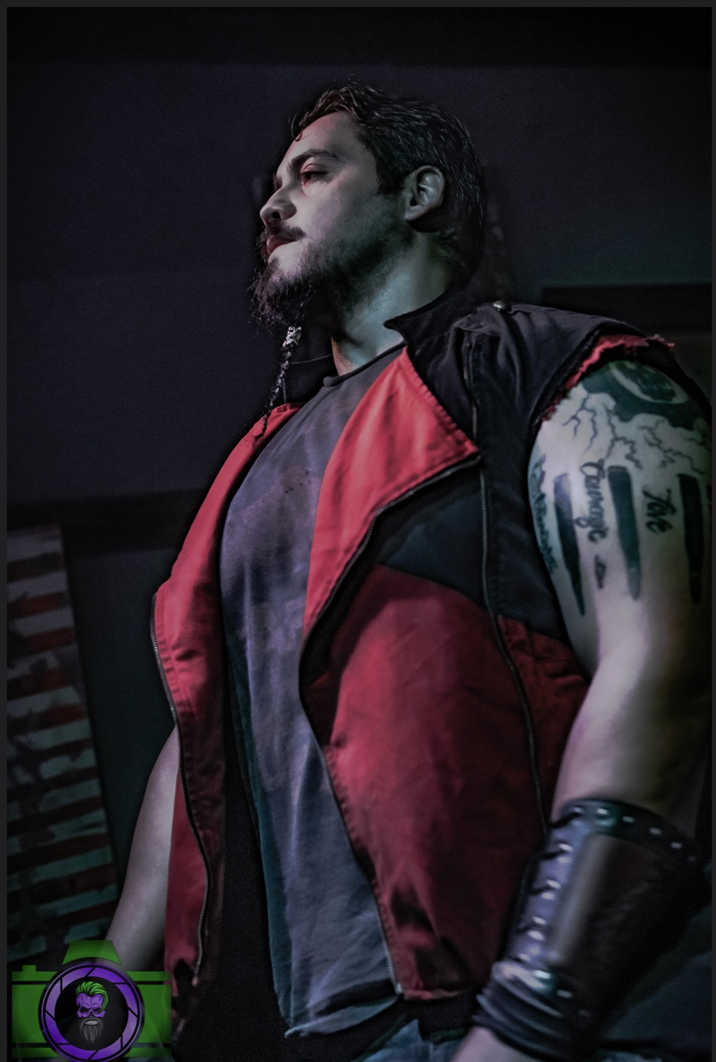Person with dark hair and beard wearing a red and black sleeveless jacket, looking to the side, with tattoos visible on arm.