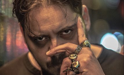 Man with dark hair and beard, holding hand with rings near face, in a dimly lit urban setting at night.