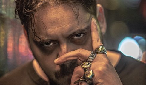 Man with dark hair and beard, holding hand with rings near face, in a dimly lit urban setting at night.