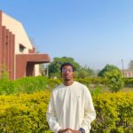 Young man standing outdoors in front of green bushes and a building, wearing a white sweatshirt and glasses.
