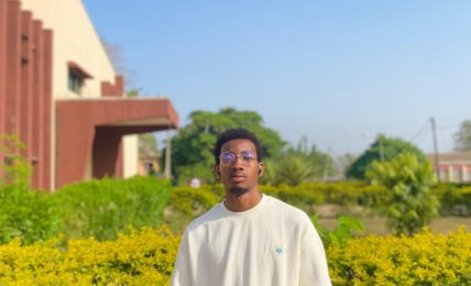Young man standing outdoors in front of green bushes and a building, wearing a white sweatshirt and glasses.
