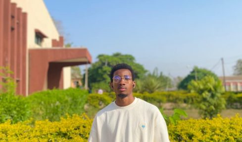 Young man standing outdoors in front of green bushes and a building, wearing a white sweatshirt and glasses.