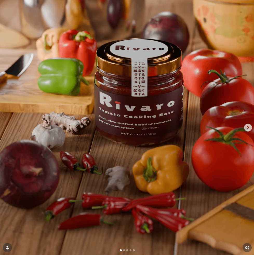 Jar of tomato cooking base surrounded by tomatoes, peppers, garlic, and chili peppers on a wooden surface.