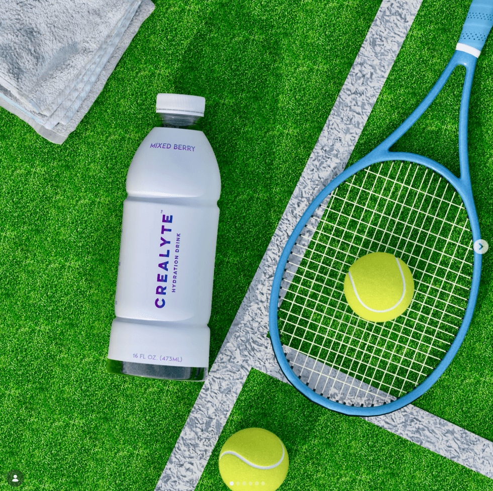 Tennis racket, tennis balls, and a bottle of berry-flavored drink on a tennis court surface.