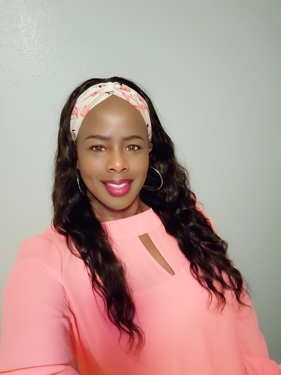 Woman with long wavy hair wearing a pink top and floral headband, smiling, against a plain background.
